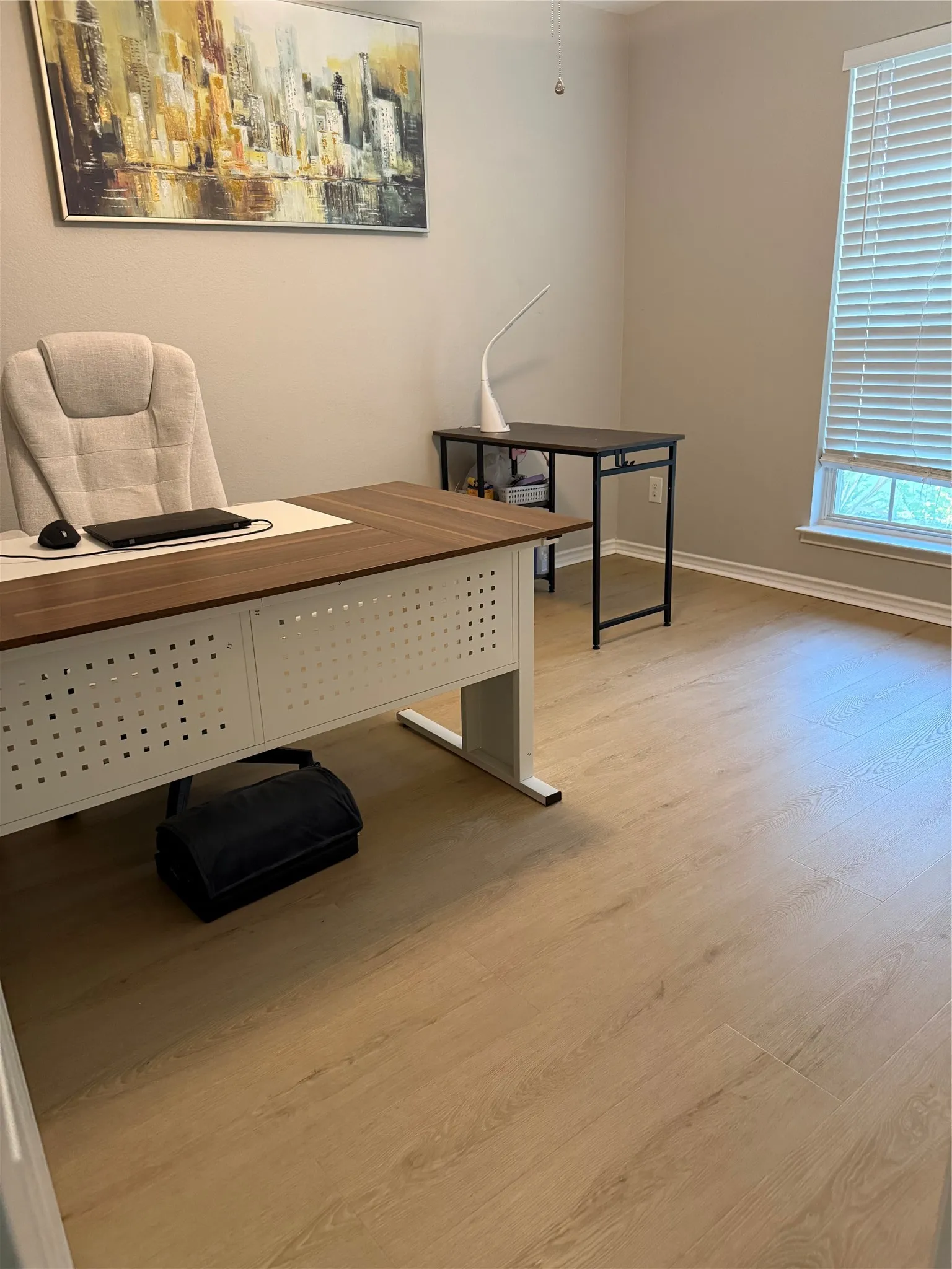 Modern office space featuring a wood-finish desk with integrated storage base, ergonomic task chair, wood-finish flooring throughout, and abundant natural light from floor-to-ceiling windows with operable blinds