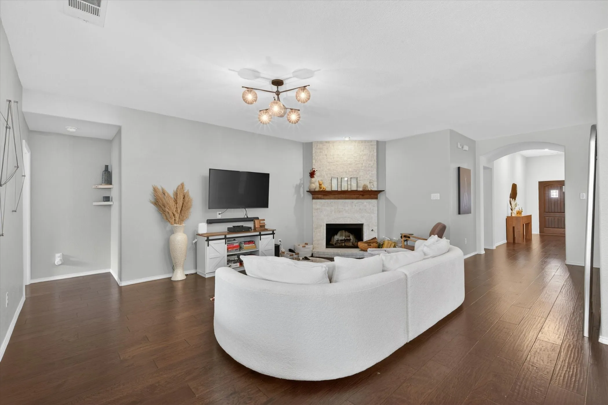 Spacious open-concept living area showcasing a curved white sectional sofa, dark wood-finish flooring throughout, and a gas fireplace with stone surround and mantel