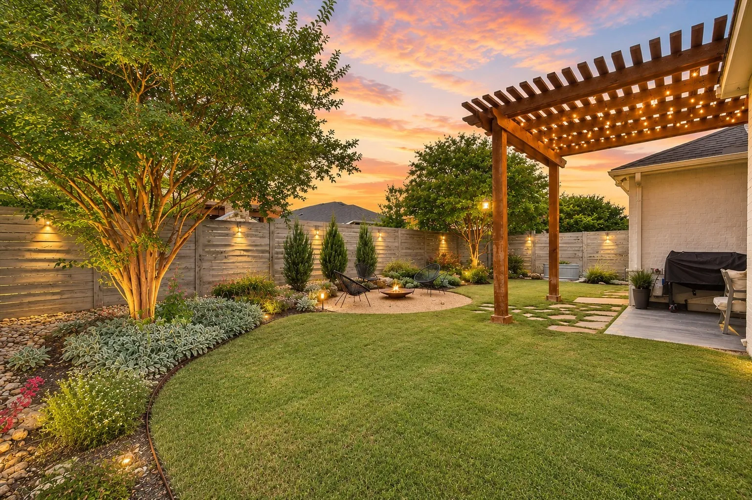 Expansive backyard oasis featuring a manicured lawn, mature specimen trees with uplighting, composite pergola with integrated shade louvers, curved landscape bed with ornamental plantings, horizontal wood-finish privacy fencing, and a covered patio area with concrete flooring ideal for entertaining