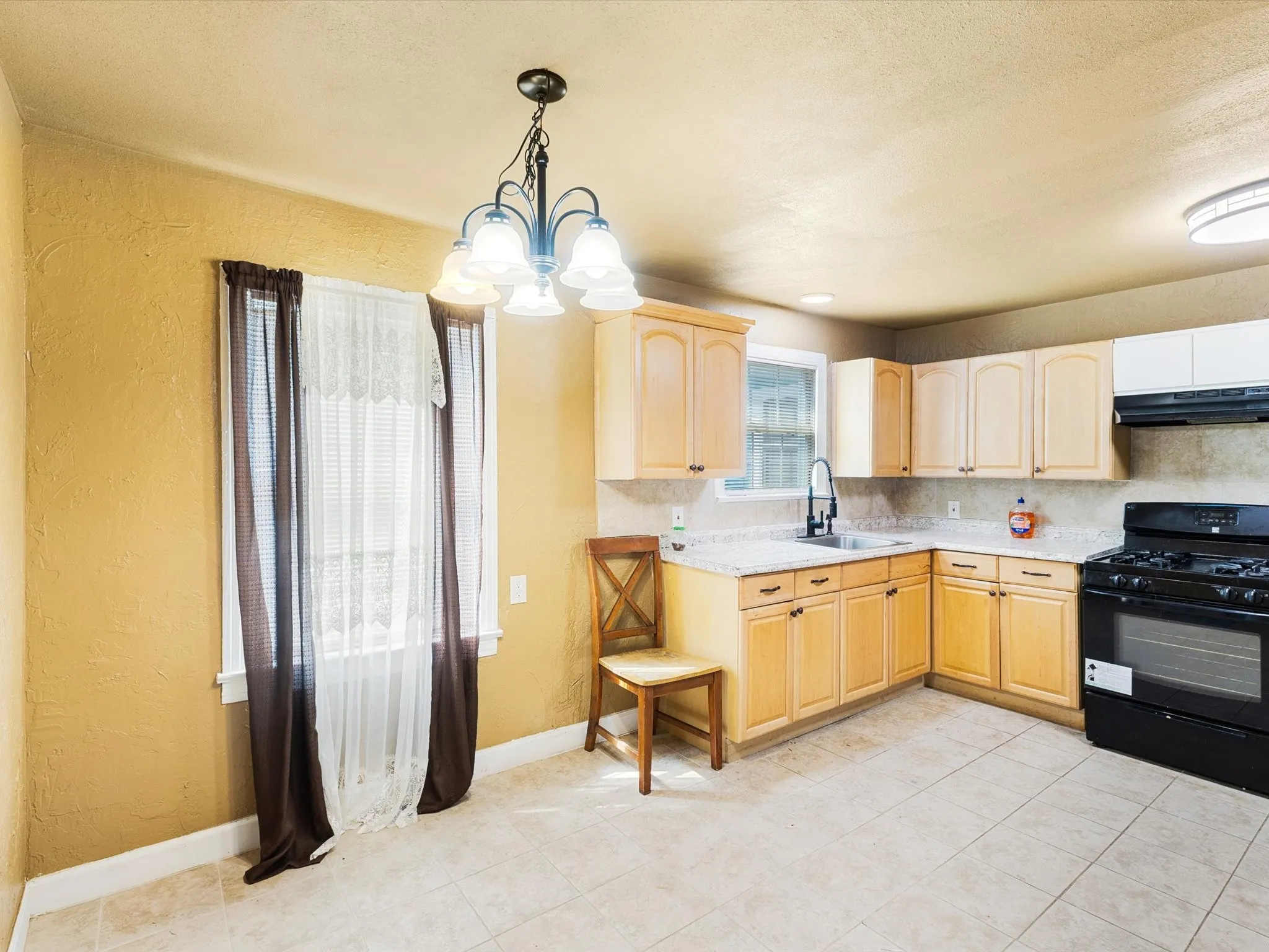 Bright and spacious kitchen featuring warm wood-finish cabinetry, black gas range with multiple burners, tile flooring, and abundant natural light from dual windows