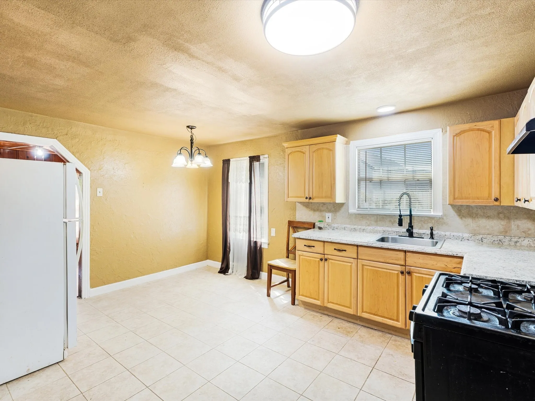 Bright and spacious kitchen featuring warm wood cabinetry, tile flooring, granite-style countertops, a gas cooktop with black range hood, and stainless steel sink with faucet