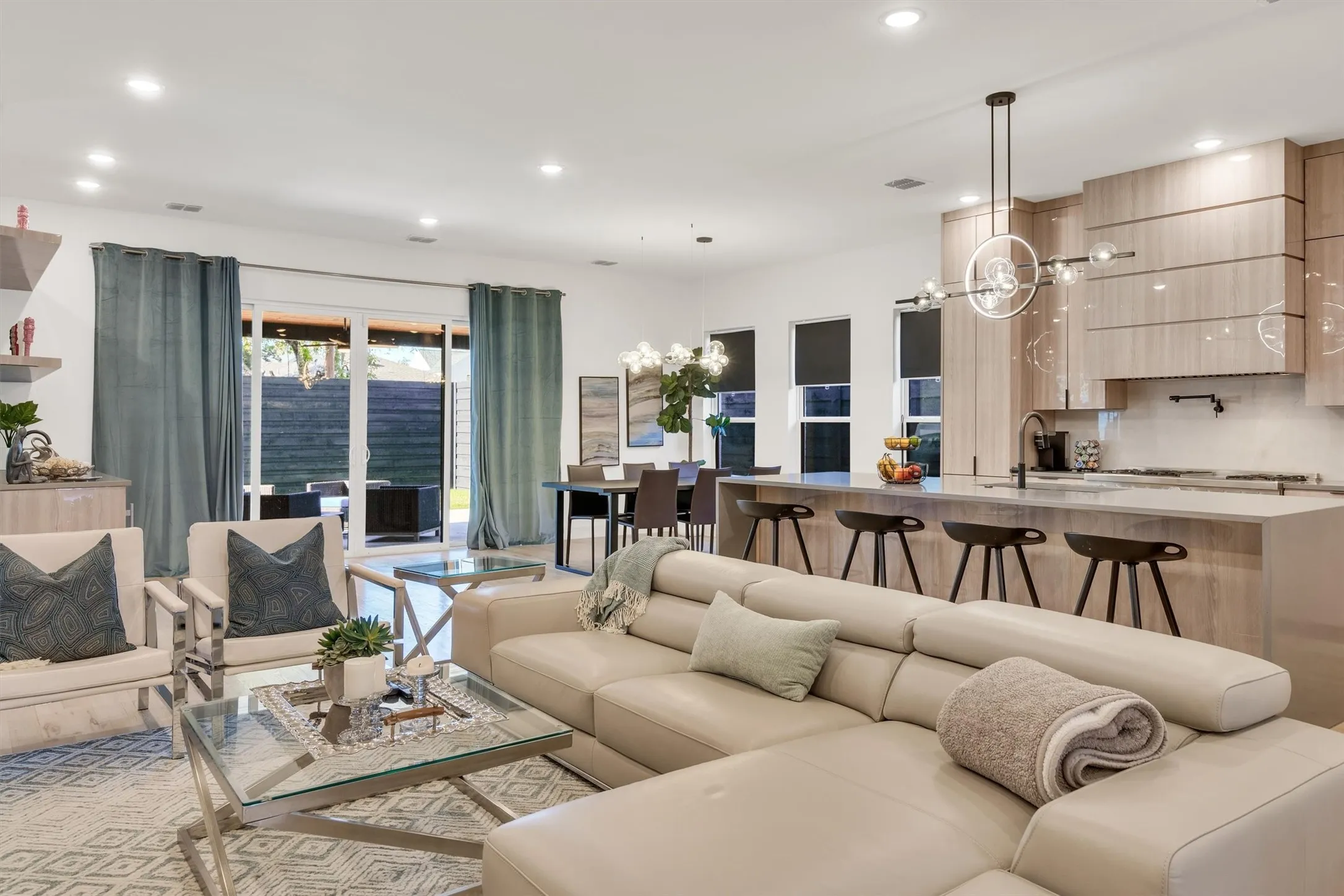 Open-concept living and dining area showcasing a curved cream sectional sofa, light wood-finish cabinetry and kitchen island with seating for three, stainless steel appliances, pendant lighting, recessed ceiling lights, and expansive windows with green curtains framing evening views