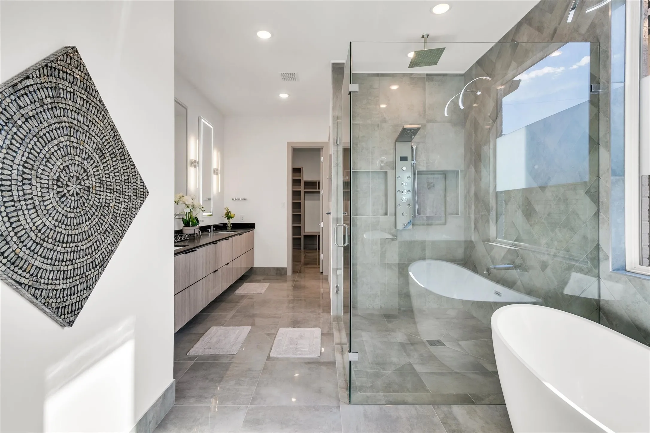 Dual vanity with dark countertops and wood-finish cabinetry, frameless glass shower enclosure, built-in shower panel system, freestanding soaking tub, and tile flooring