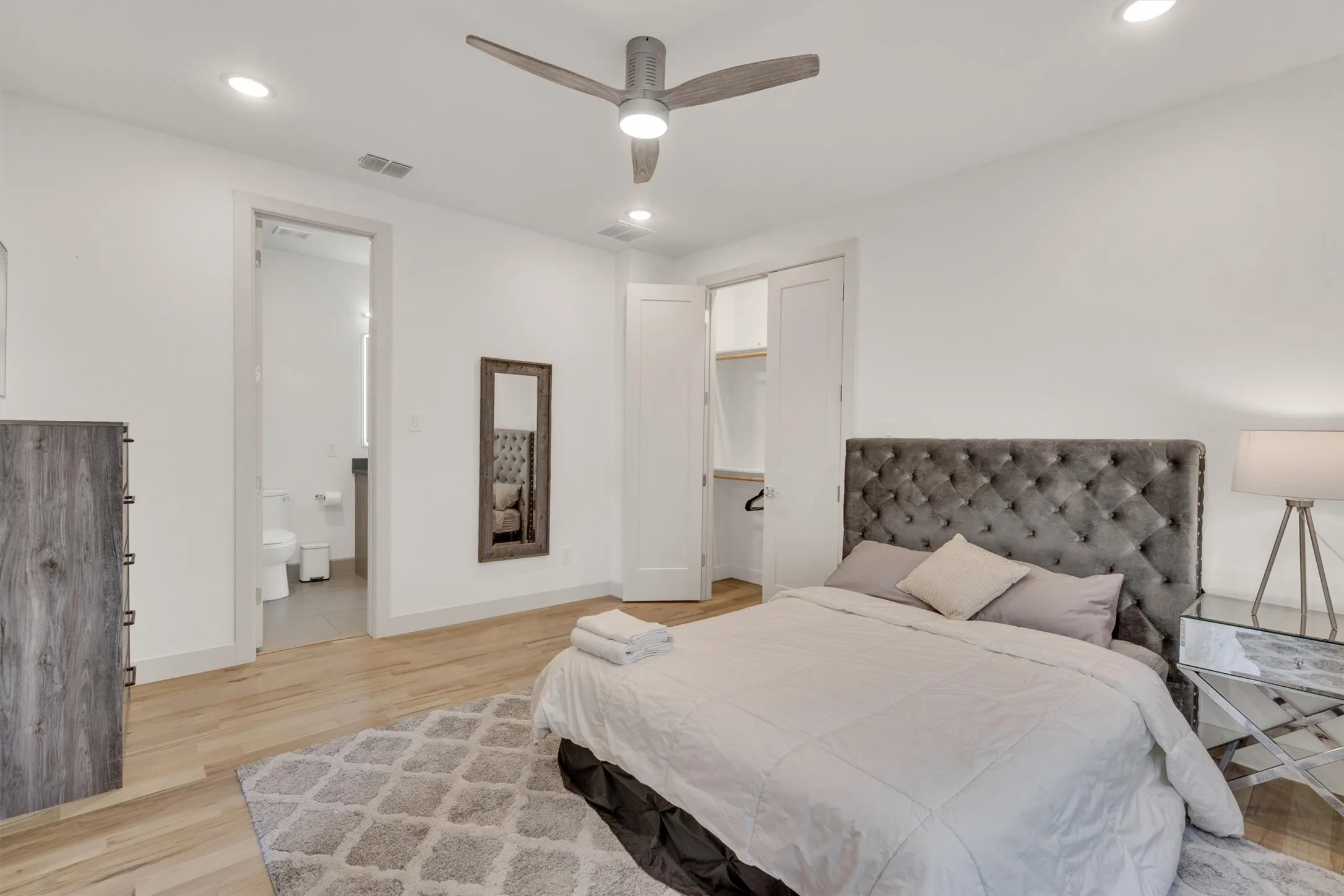 Spacious primary bedroom showcasing a tufted upholstered headboard, wood-finish flooring throughout, recessed ceiling lighting, and a ceiling fan