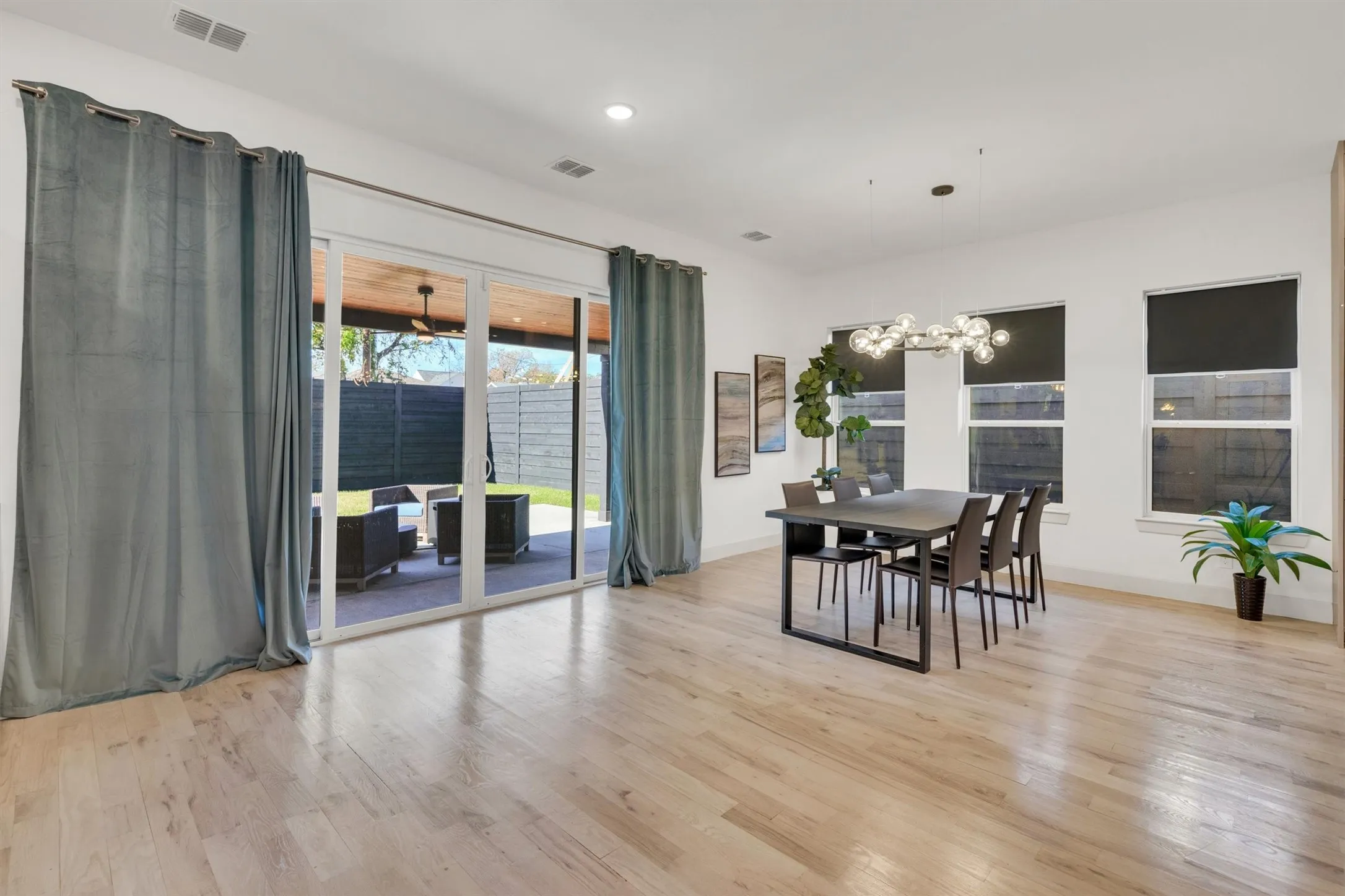 Open-concept living area showcasing gleaming wood-finish flooring throughout, expansive glass sliding doors with transom windows opening to an outdoor patio, custom gray drapery panels, recessed ceiling lighting, a dining table with seating for multiple guests, and abundant natural light from multiple windows with integrated shading systems
