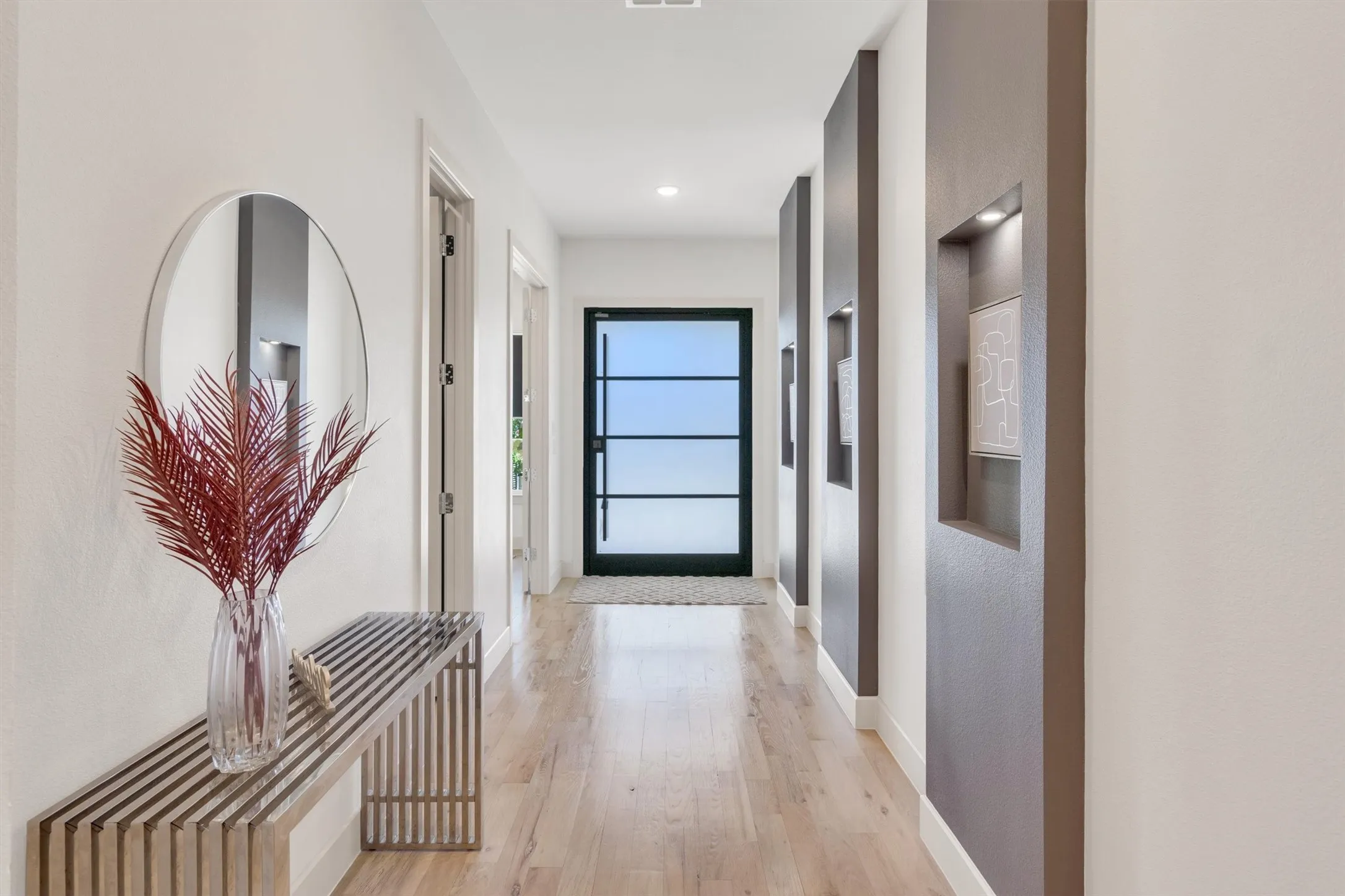 Light wood-finish flooring extending through the hallway, modern dark-framed entry door with frosted glass panels, recessed ceiling lighting, and built-in wall niches with integrated illumination