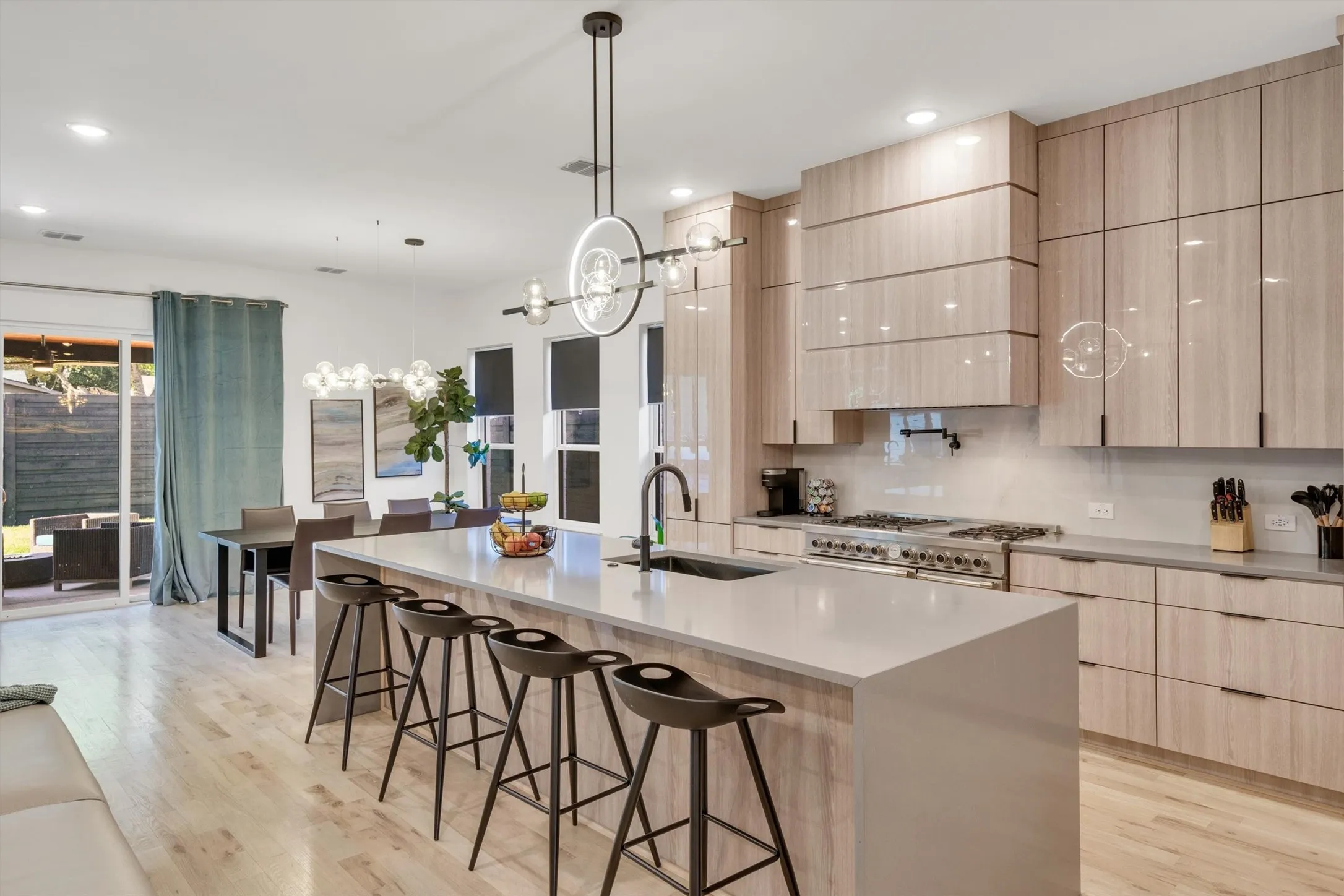 Modern kitchen featuring light wood-finish cabinetry with glass-front upper cabinets, a large waterfall-edge quartz island with seating for multiple guests, stainless steel gas cooktop and range hood, and pendant lighting
