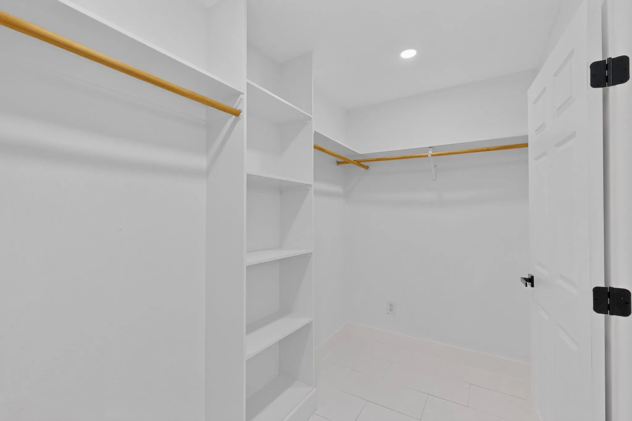 Spacious walk-in closet featuring built-in shelving, dual hanging rods with brass finish hardware, recessed ceiling lighting, and frameless glass entry door with matte black hardware