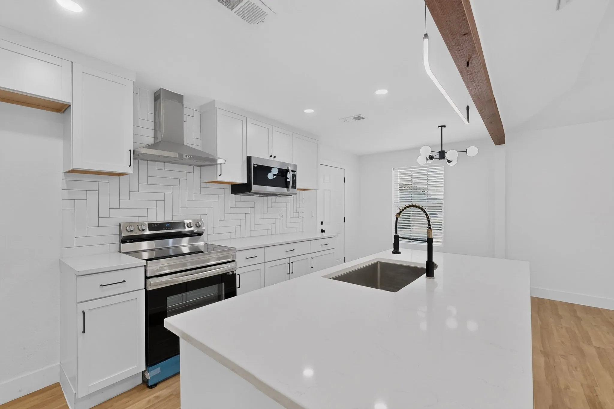 Modern kitchen featuring white cabinetry, stainless steel appliances including a gas range and range hood, white subway tile backsplash, quartz countertops with waterfall edge, undermount sink with contemporary faucet, and wood-finish flooring throughout