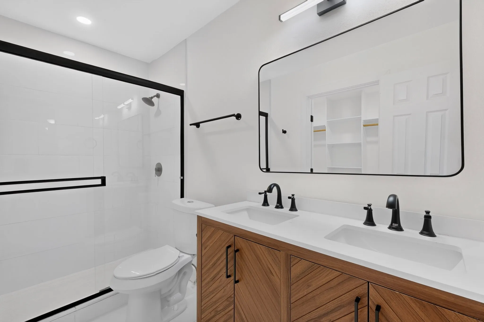 Spa-inspired primary bathroom featuring a dual-sink vanity with warm wood-finish cabinetry, white quartz countertops, and matte black hardware and faucets