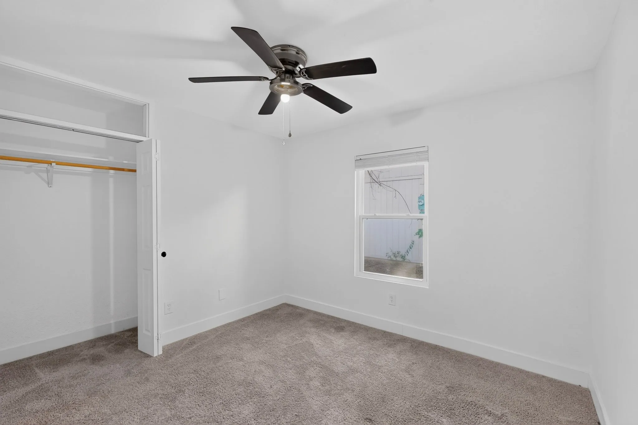 Bright secondary bedroom featuring neutral carpeting, ceiling fan with integrated lighting, spacious closet with wooden rod, and large window allowing natural light
