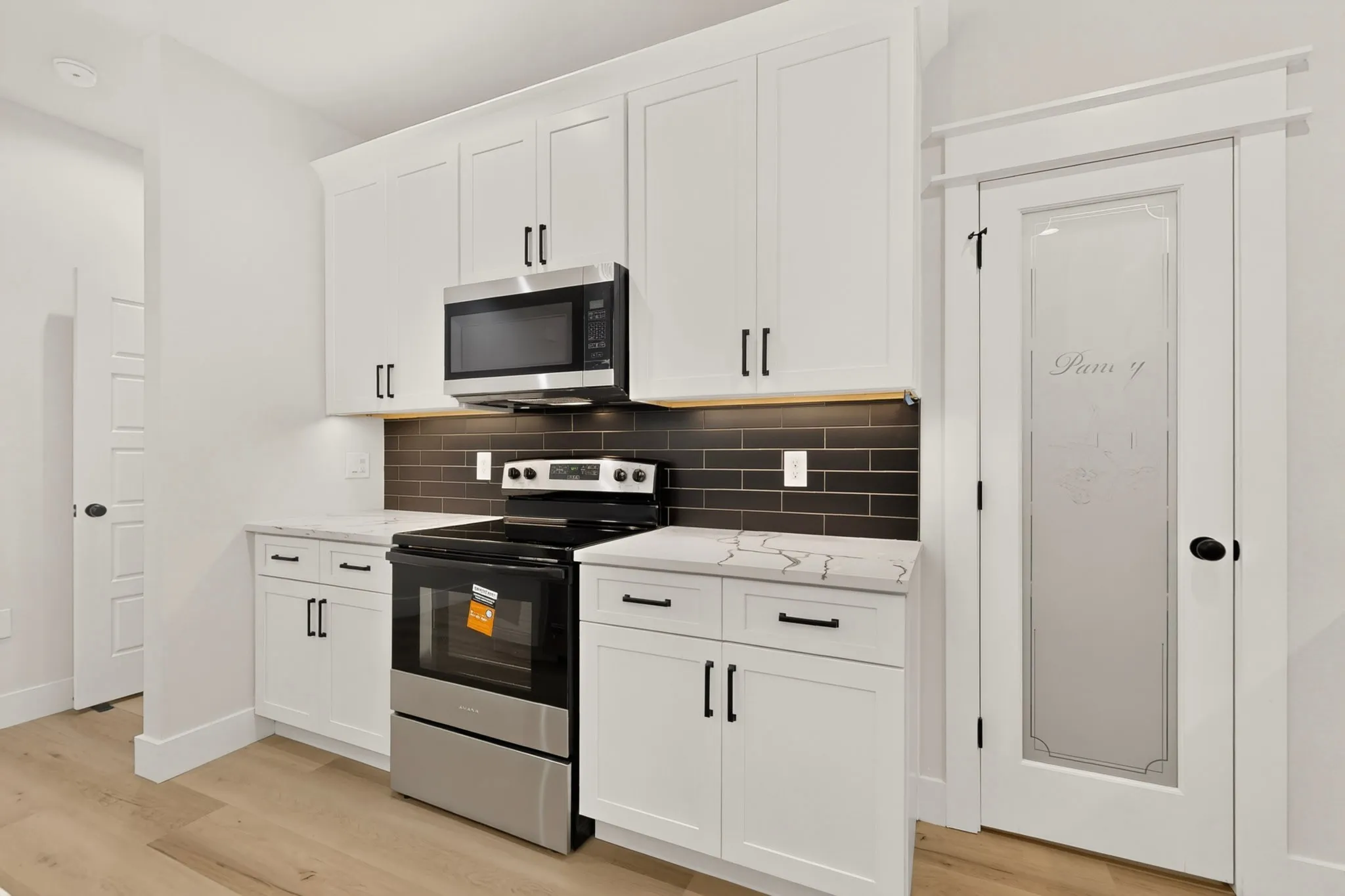 White shaker cabinetry with black hardware, stainless steel appliances, dark subway tile backsplash, light wood-finish flooring, and a frosted glass pantry door