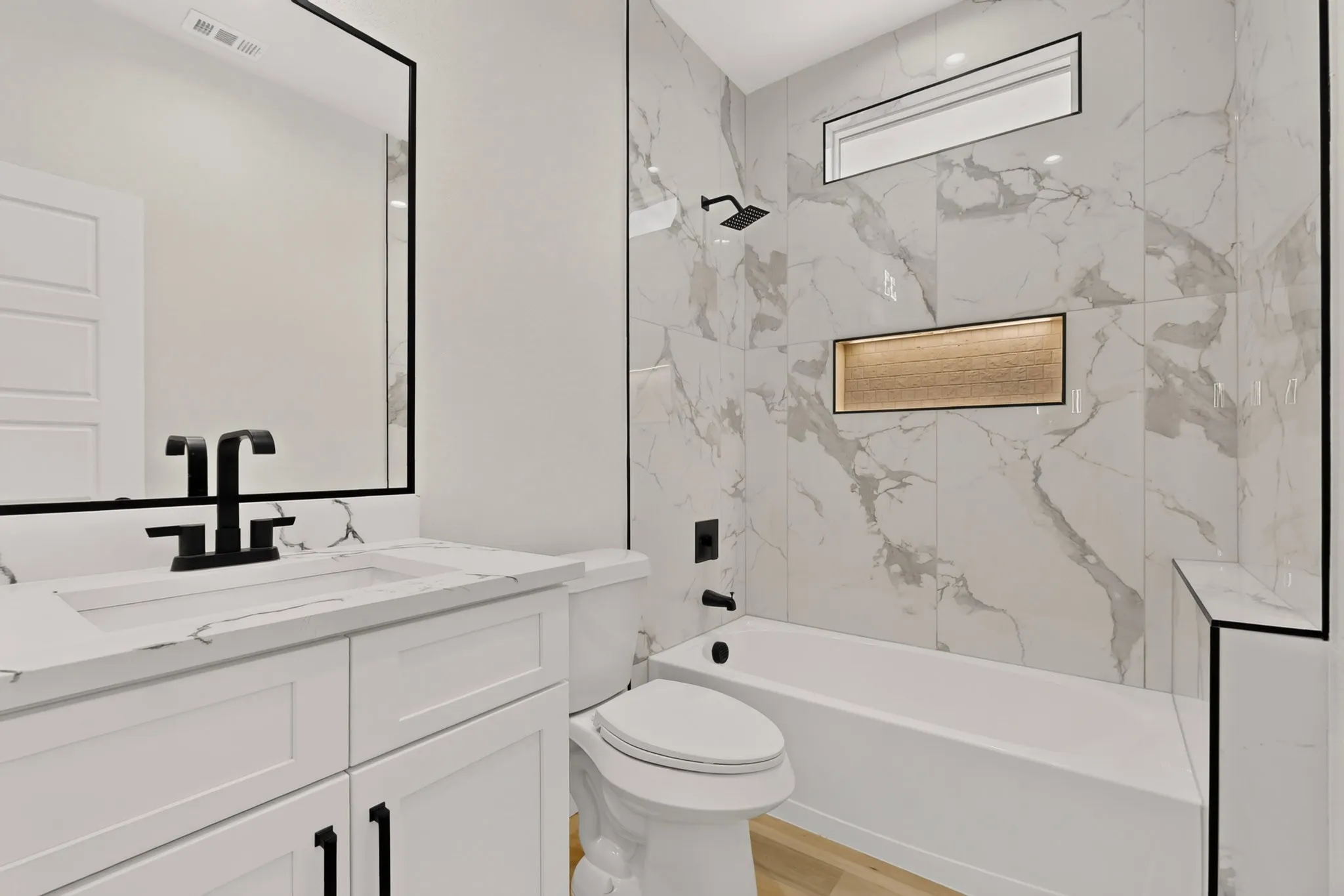 White vanity with integrated sink and matte black faucet, framed mirror, full-height wall tiling, built-in bathtub, and wall niche with textured accent