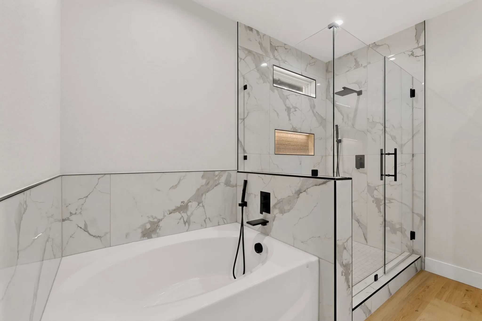 Freestanding soaking tub with matte black fixtures, glass-enclosed shower with a rainfall showerhead, and a tiled accent niche