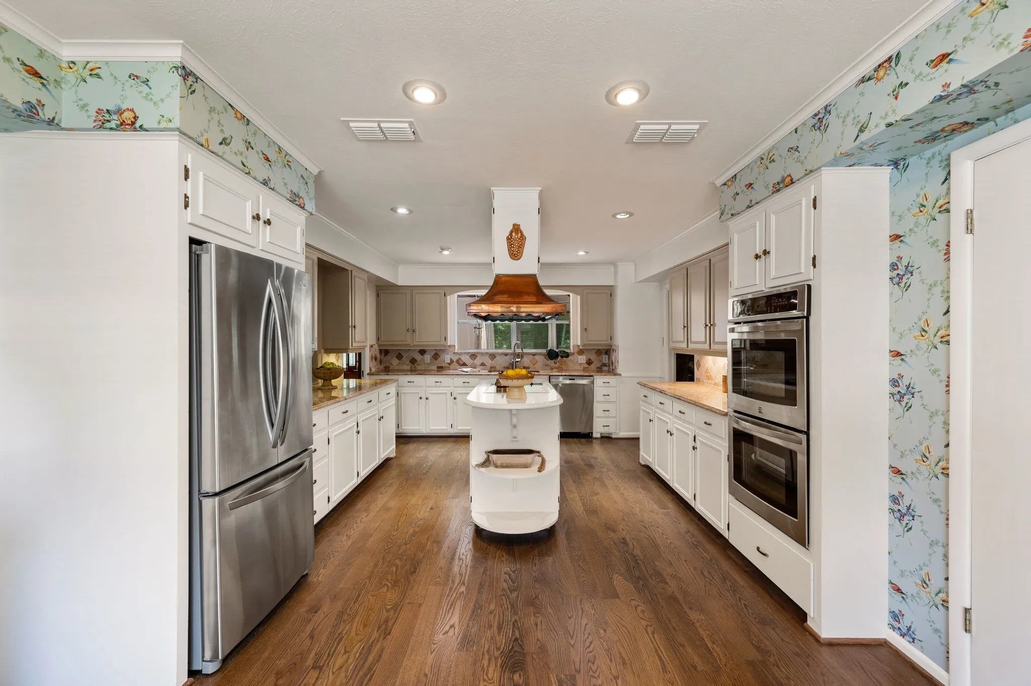 Beautiful copper vent hood takes center stage in this pretty kitchen with wallpaper, stainless appliances and solid oak floors