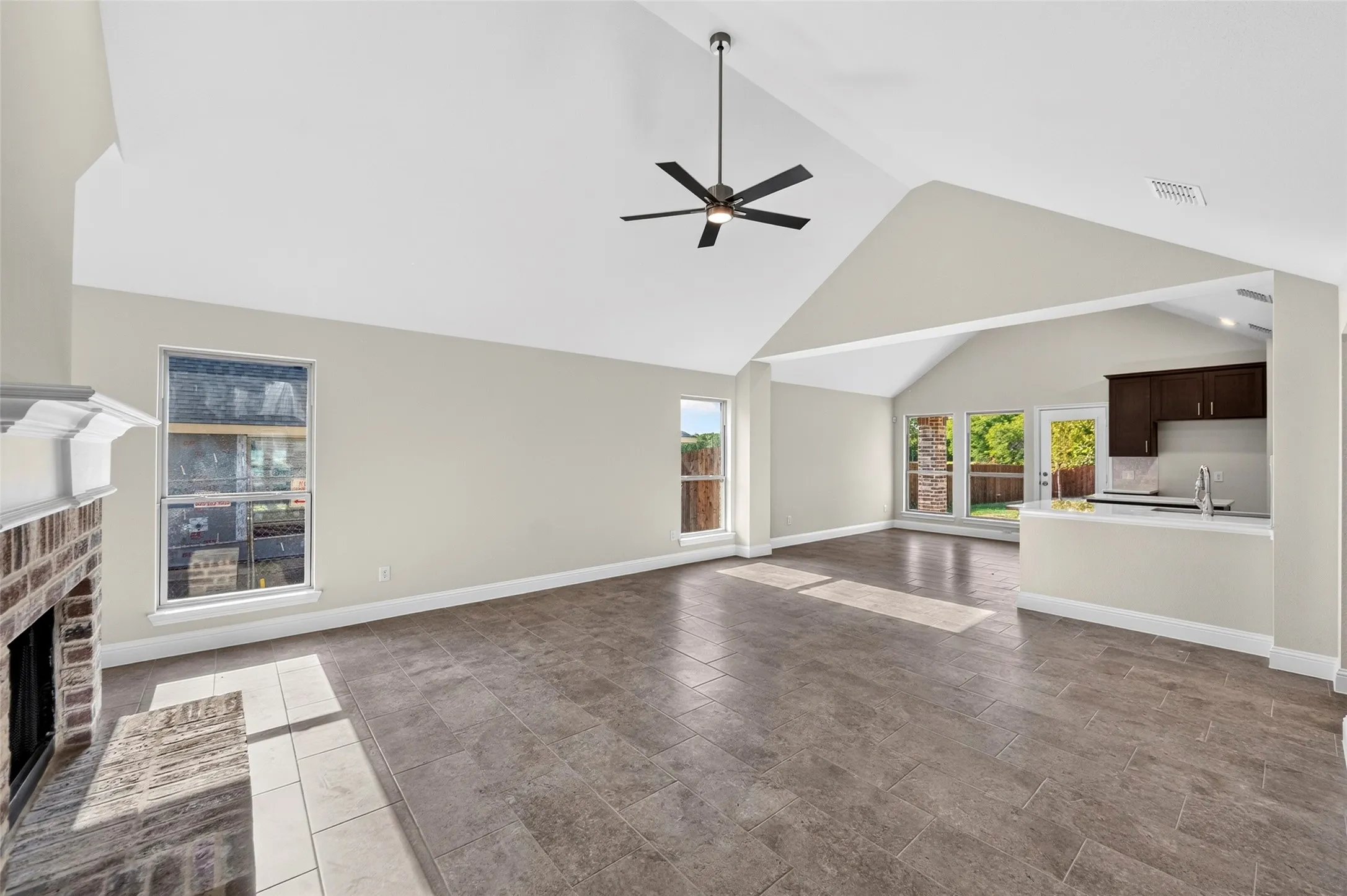 Vaulted ceiling with a ceiling fan, brick fireplace, tile flooring, multiple windows, and a peninsula kitchen