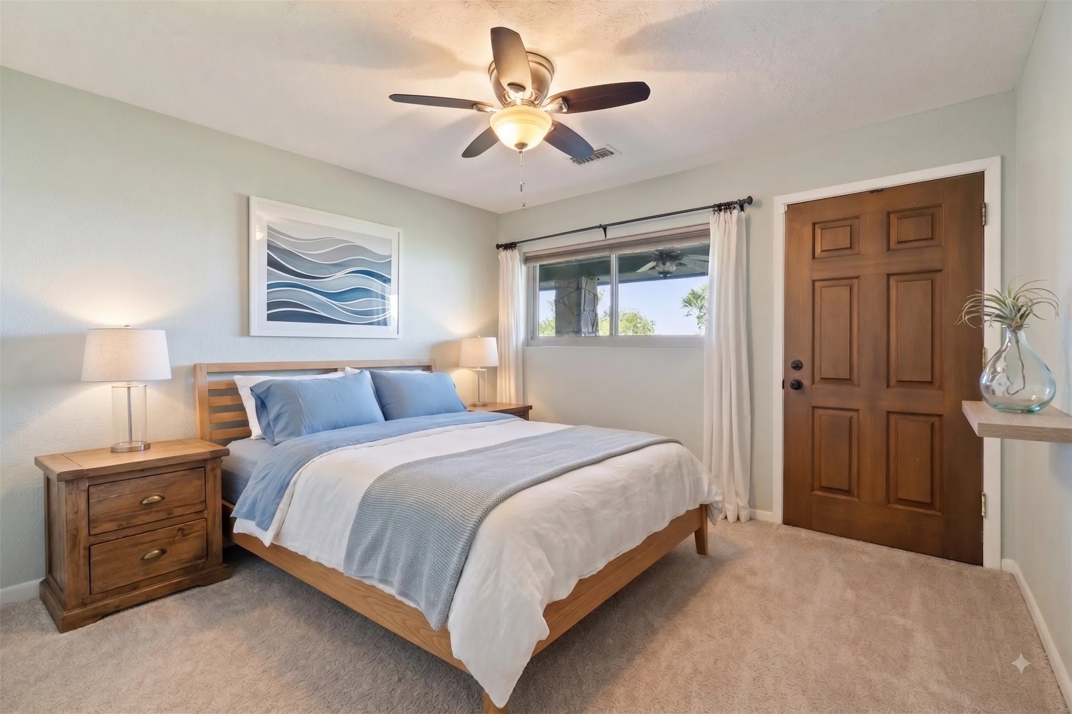 Serene primary bedroom showcasing warm wood-finish furnishings, soft blue and neutral bedding, carpeted flooring, and abundant natural light from large windows