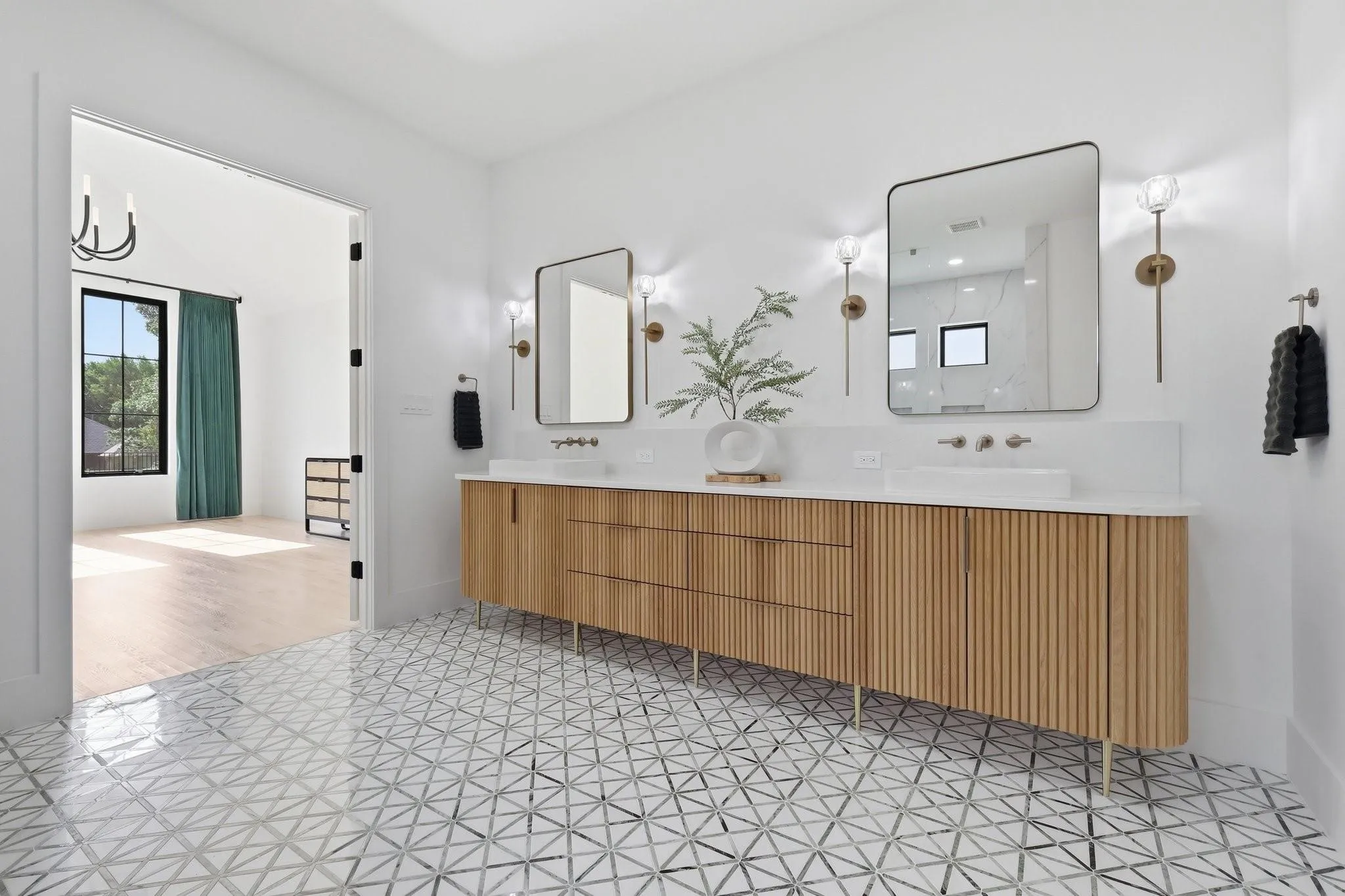 The Ensuite features a quartz vanity with fluted white oak cabinetry by Hunter Trim & Cabinets, dual brass wall-mount faucets, brushed gold sconce lighting, and designer geometric marble tile flooring.
