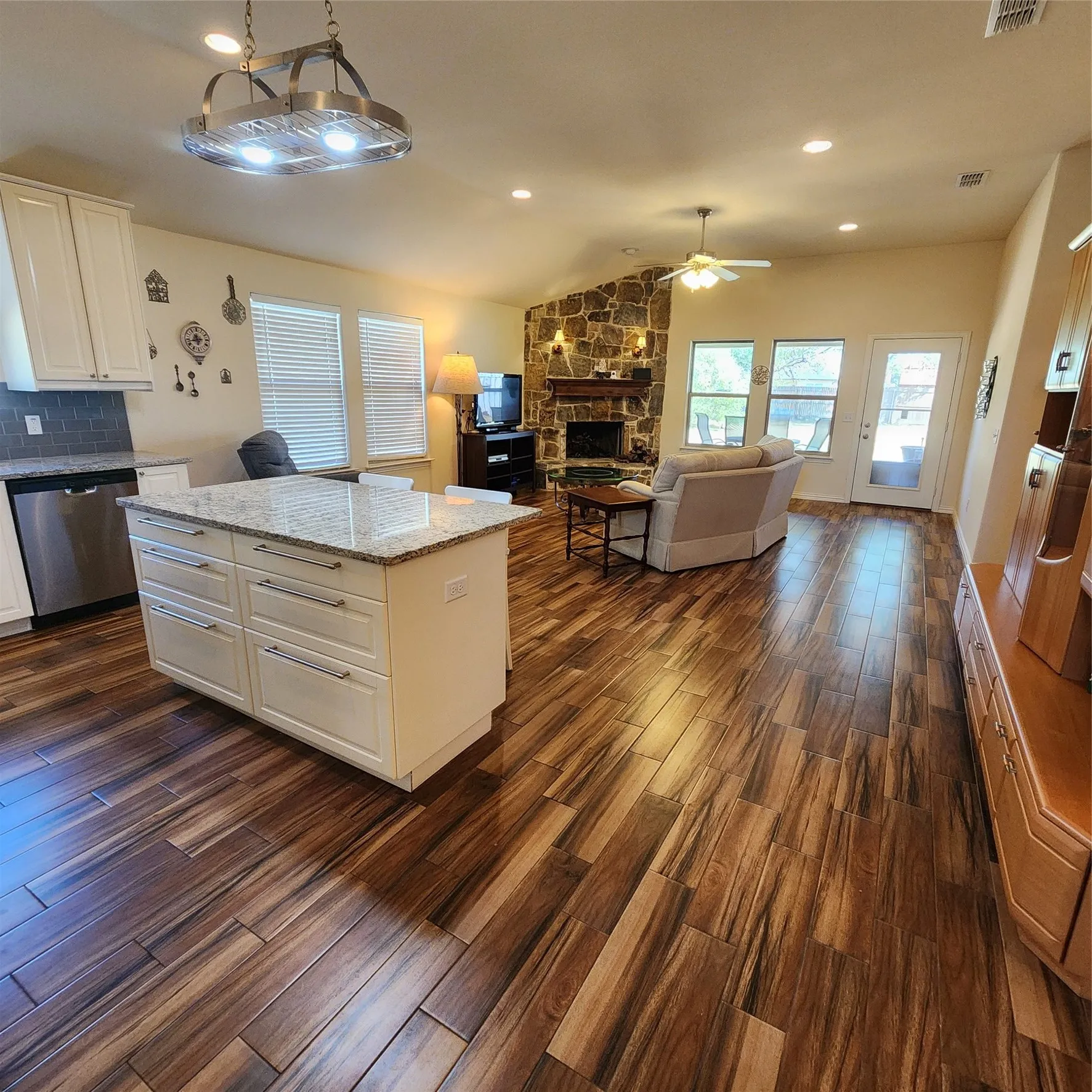 Open-concept living space with wood-finish flooring, a stone fireplace, and a kitchen island featuring a light-toned countertop