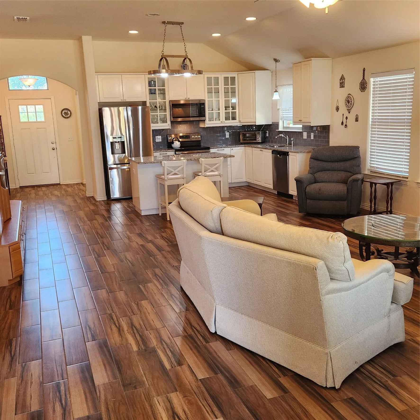 Open-concept living space featuring wood-finish flooring, vaulted ceilings, and a kitchen with white cabinetry, stainless steel appliances, and a tiled backsplash