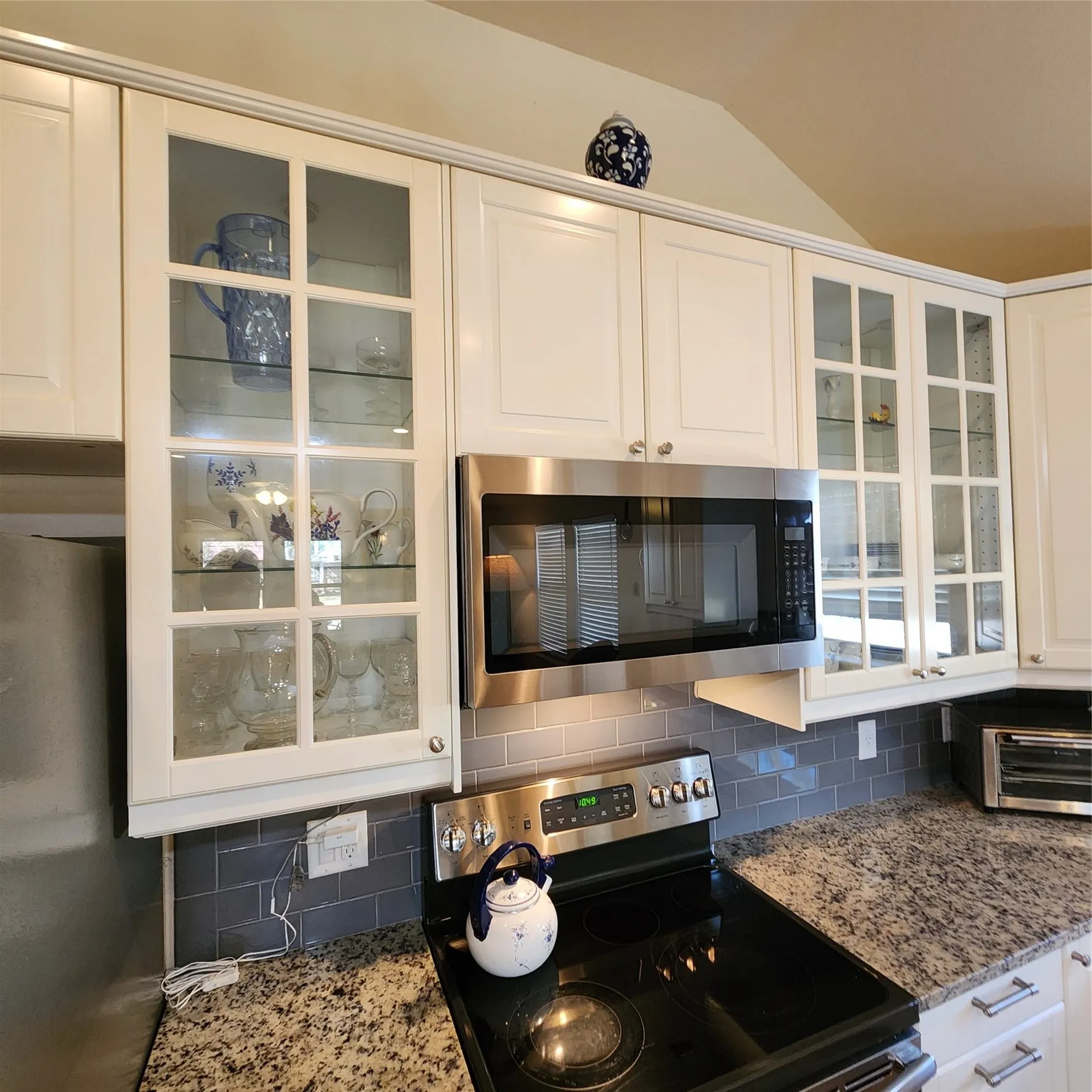 White cabinetry with glass-front display doors, stainless steel microwave, black electric range, subway tile backsplash, and speckled countertops