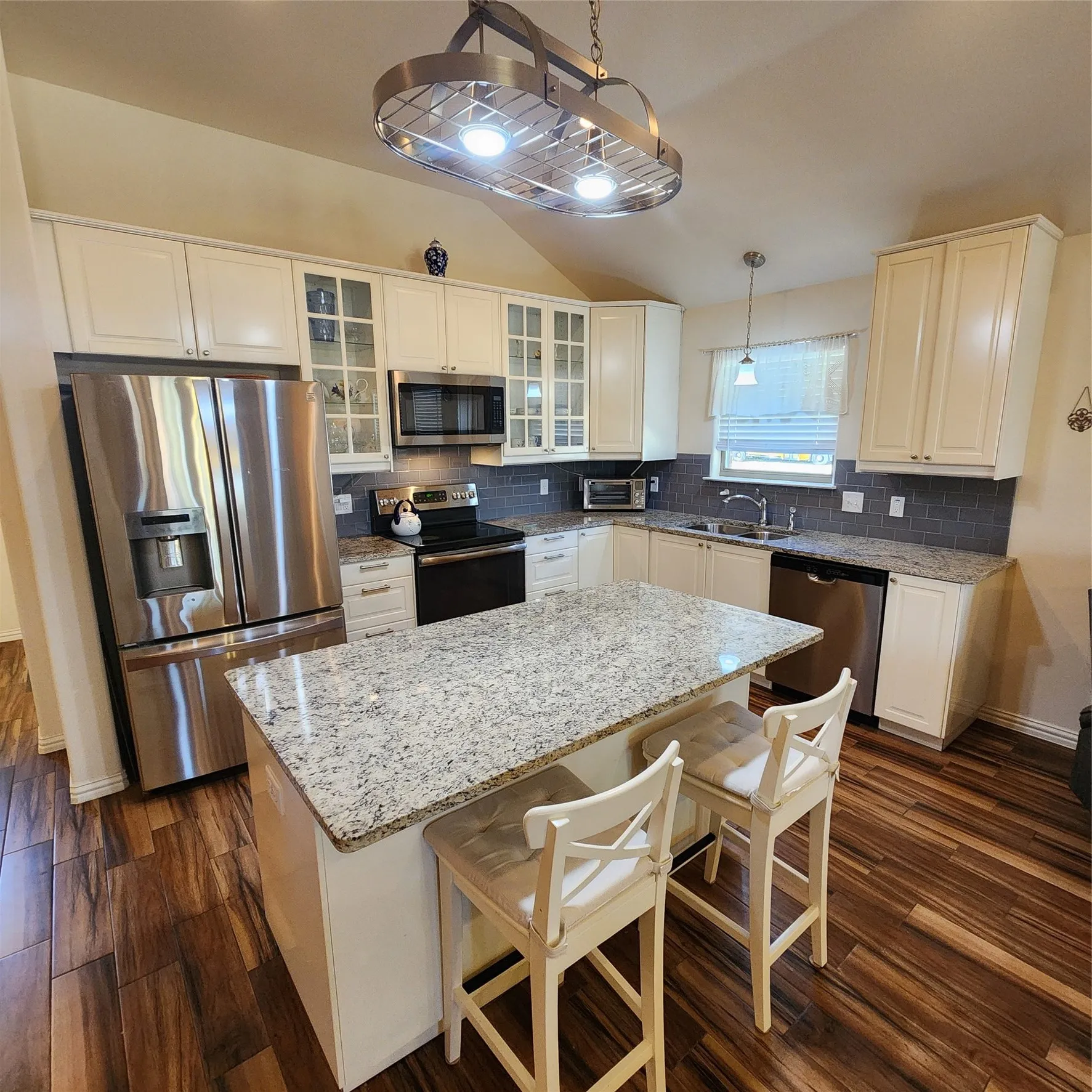 Vaulted ceiling with an overhead light fixture, wood-finish flooring, white cabinetry, a kitchen island with stone countertops, and stainless steel appliances
