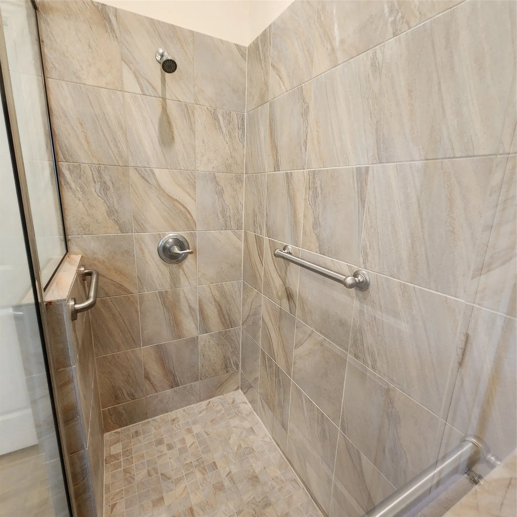 Walk-in shower featuring large format wall tiles, mosaic tile flooring, a chrome showerhead, a chrome single-handle control, and a stainless steel grab bar