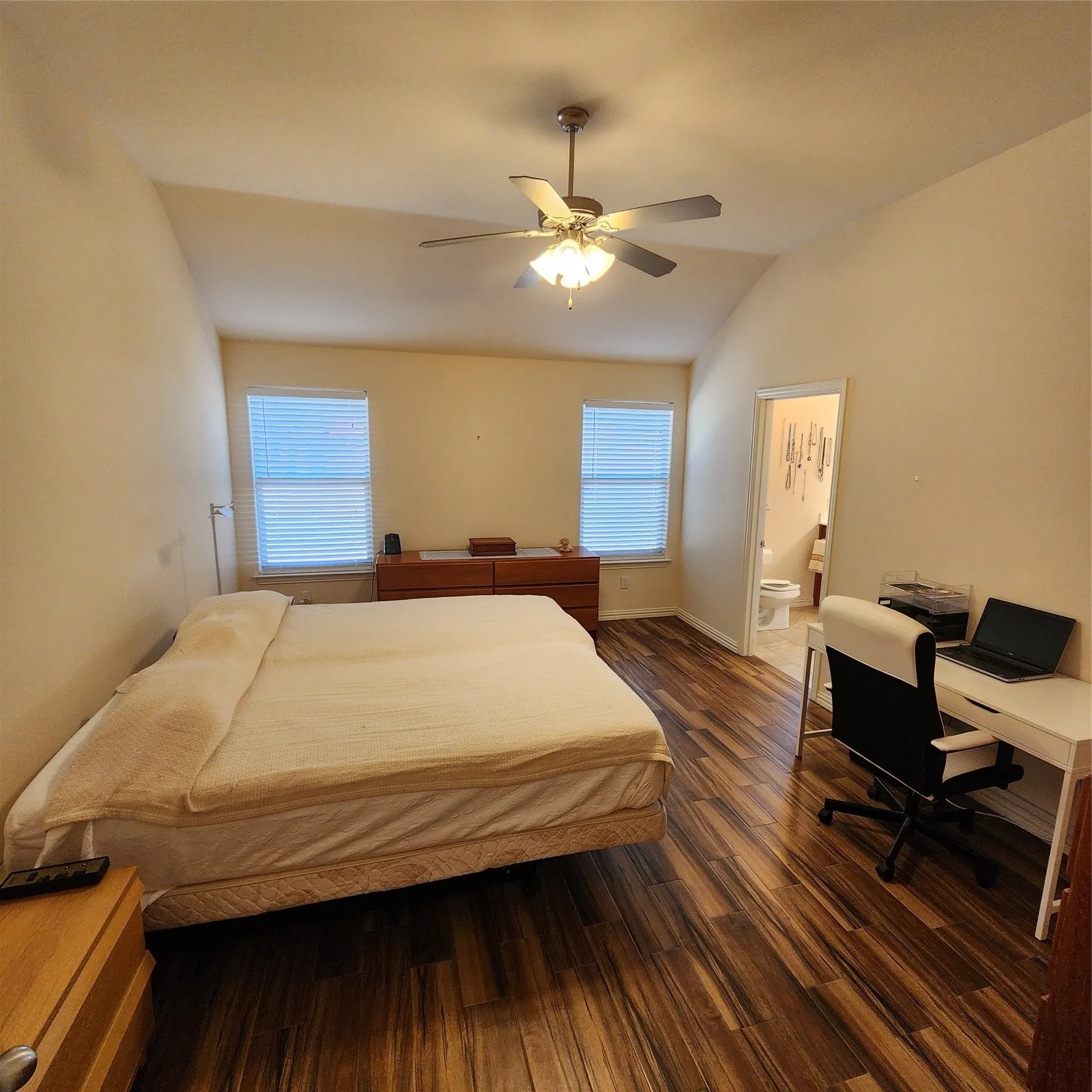 Vaulted ceiling, wood-finish flooring, two windows with blinds, ceiling fan with integrated lighting, and a doorway opening to a bathroom