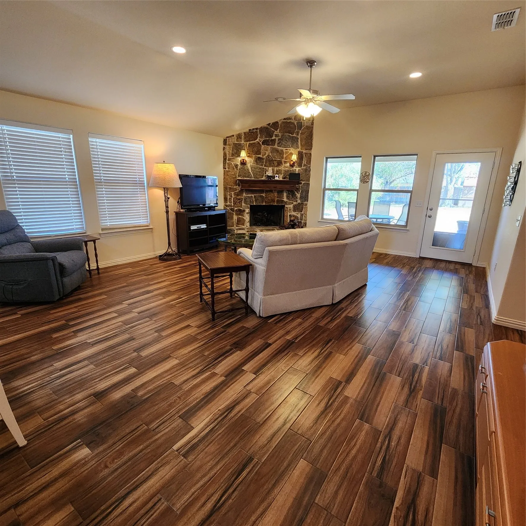 Vaulted ceiling with a ceiling fan, stone fireplace with a wood mantle, wood-finish flooring, recessed lighting, and ample natural light from multiple windows and a glass-paneled door
