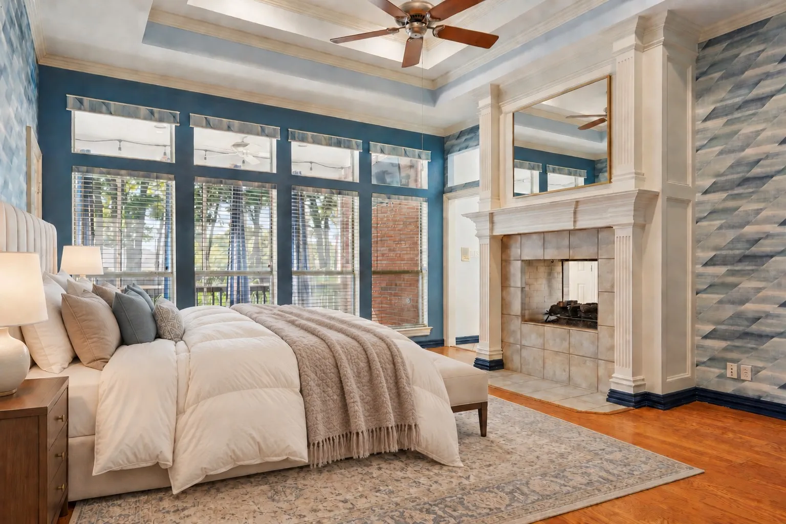 Expansive window wall, tray ceiling with wood-finish ceiling fan, hardwood flooring, fireplace with tile surround, and decorative accent wall
