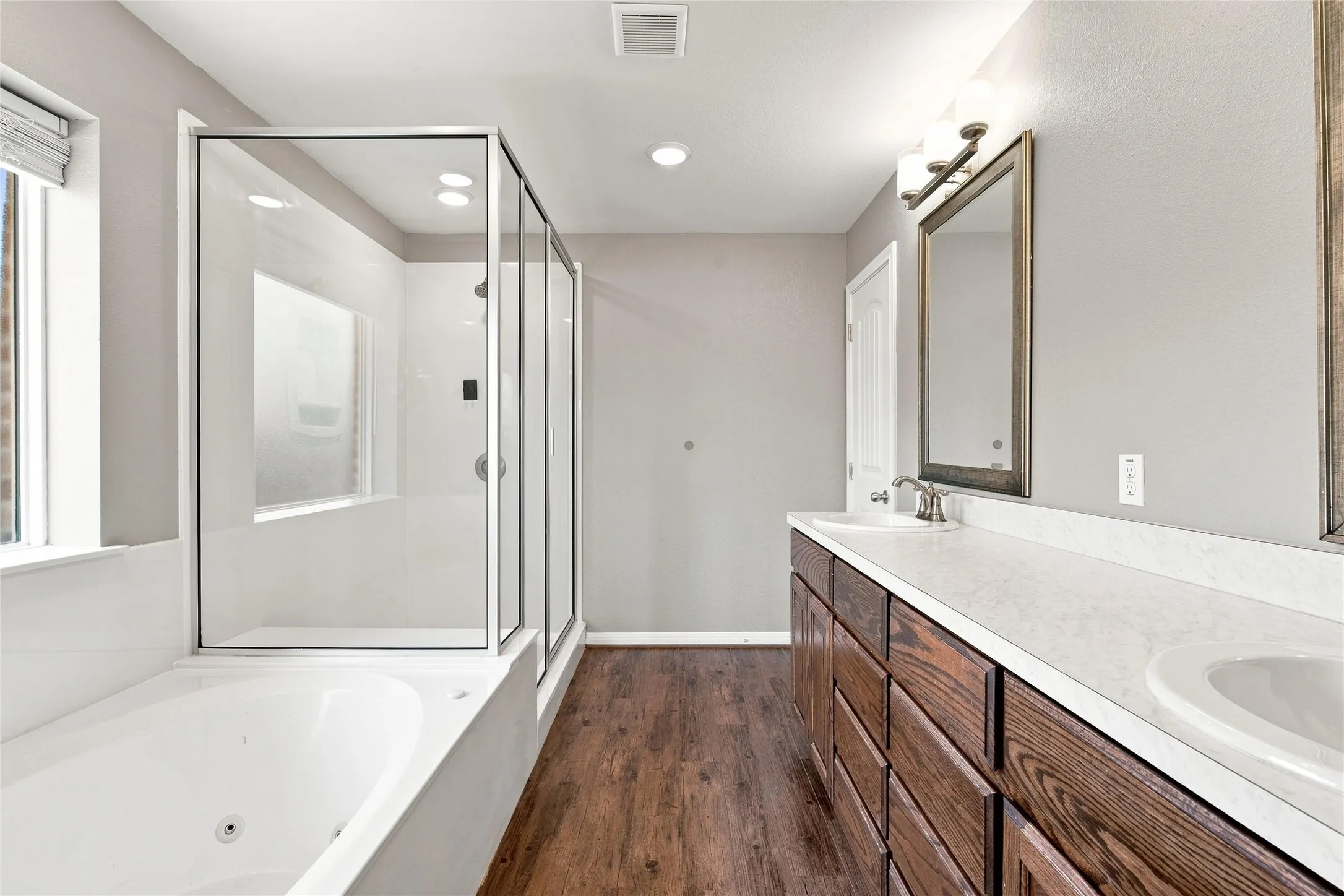 Dual vanity with integrated sinks, wood-finish cabinetry, frameless glass shower enclosure, built-in bathtub, and wood-finish flooring
