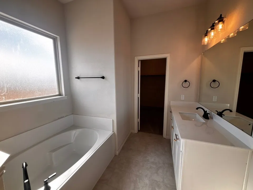 Built-in bathtub with black fixtures, expansive vanity with dual sinks, overhead lighting fixture, and tile flooring