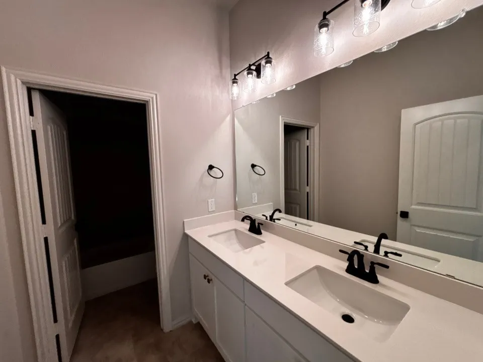 Dual undermount sinks with matte black fixtures, set into a light-toned countertop, and illuminated by a multi-bulb vanity light