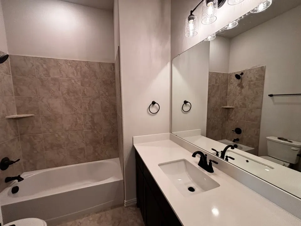 Built-in bathtub with tiled surround, solid surface vanity countertop, undermount sink, and dark-finish hardware