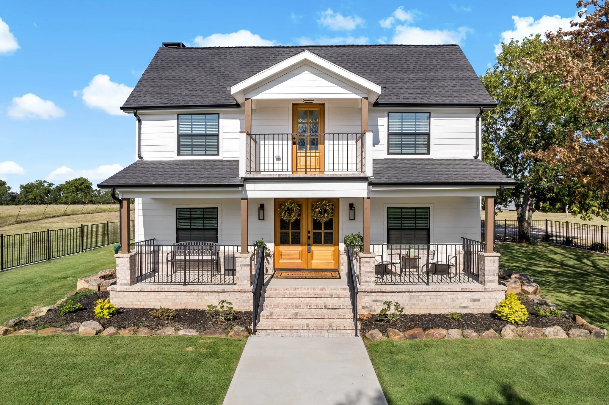 White siding exterior with a dark shingle roof, double wood front doors, a second-story balcony with metal railing, and beautiful landscaping.