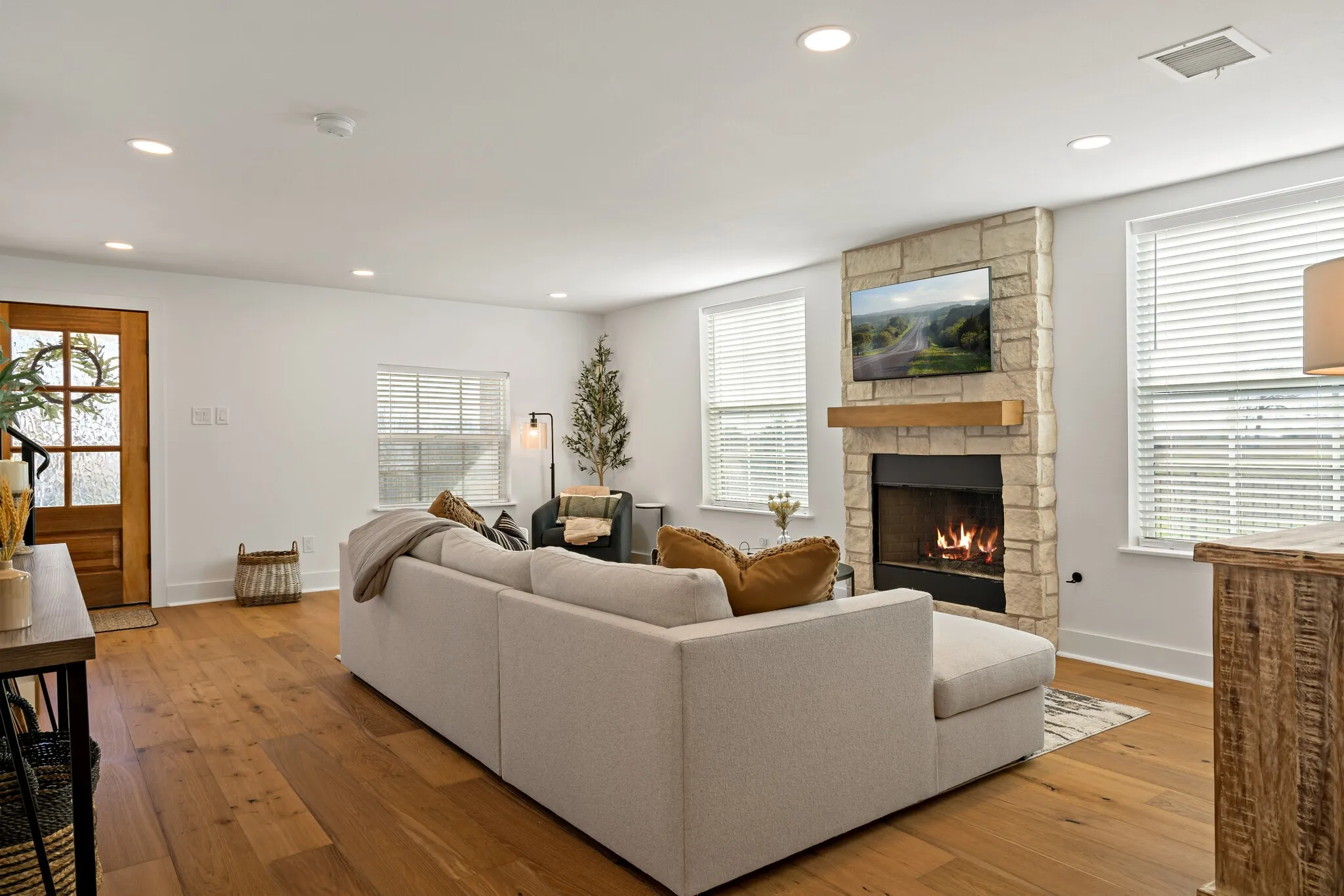 Spacious living room featuring hardwood flooring, a stone fireplace with gas insert, recessed ceiling lighting, and expansive windows with plantation shutters allowing natural light throughout the room