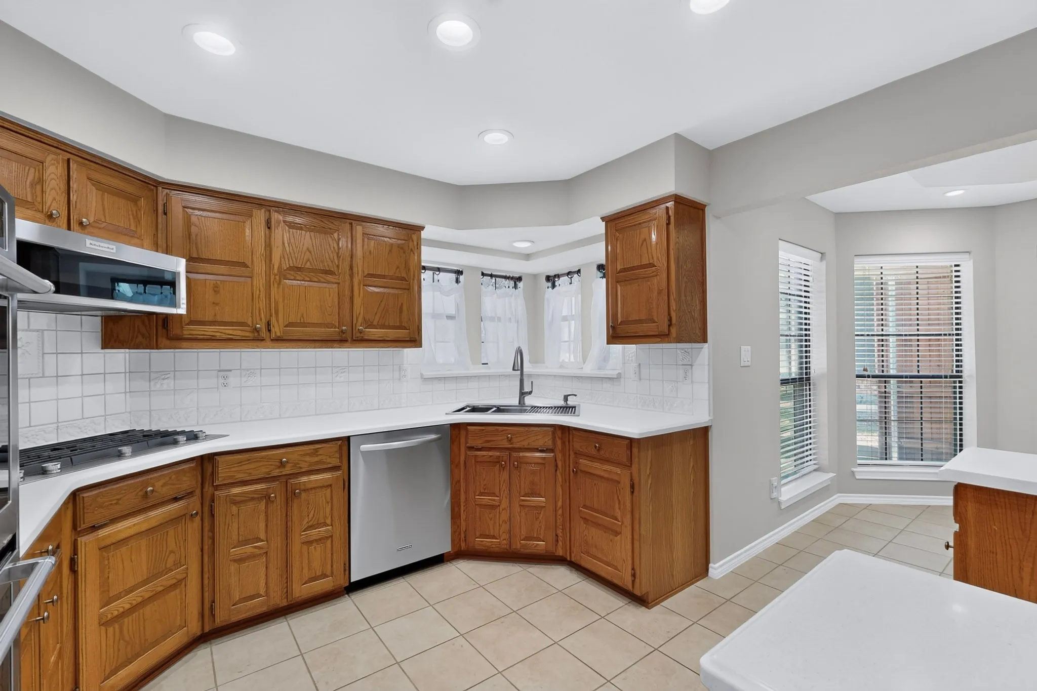 Spacious kitchen featuring warm wood cabinetry, stainless steel appliances, ceramic tile flooring, white subway tile backsplash, and a corner sink with natural light from multiple windows