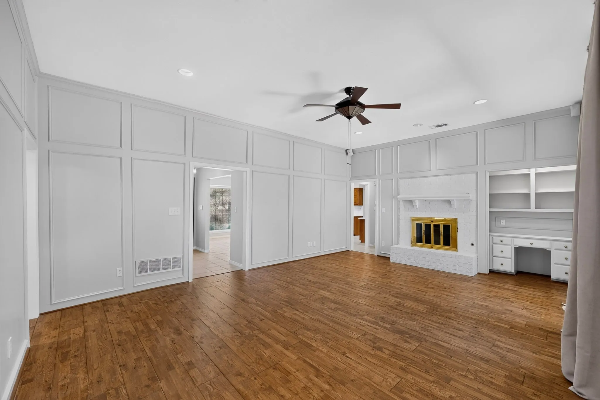 Spacious room featuring wood-finish flooring, wainscoting paneling, a brick fireplace with a brass-tone insert, built-in shelving, and a ceiling fan