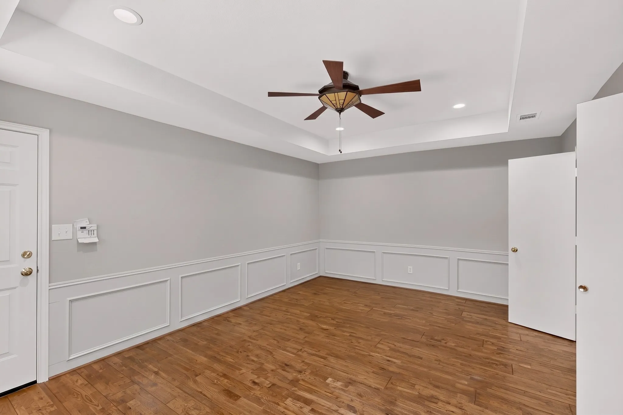 Wood-finish flooring, tray ceiling with recessed lighting, ceiling fan with integrated light fixture, and wainscoting