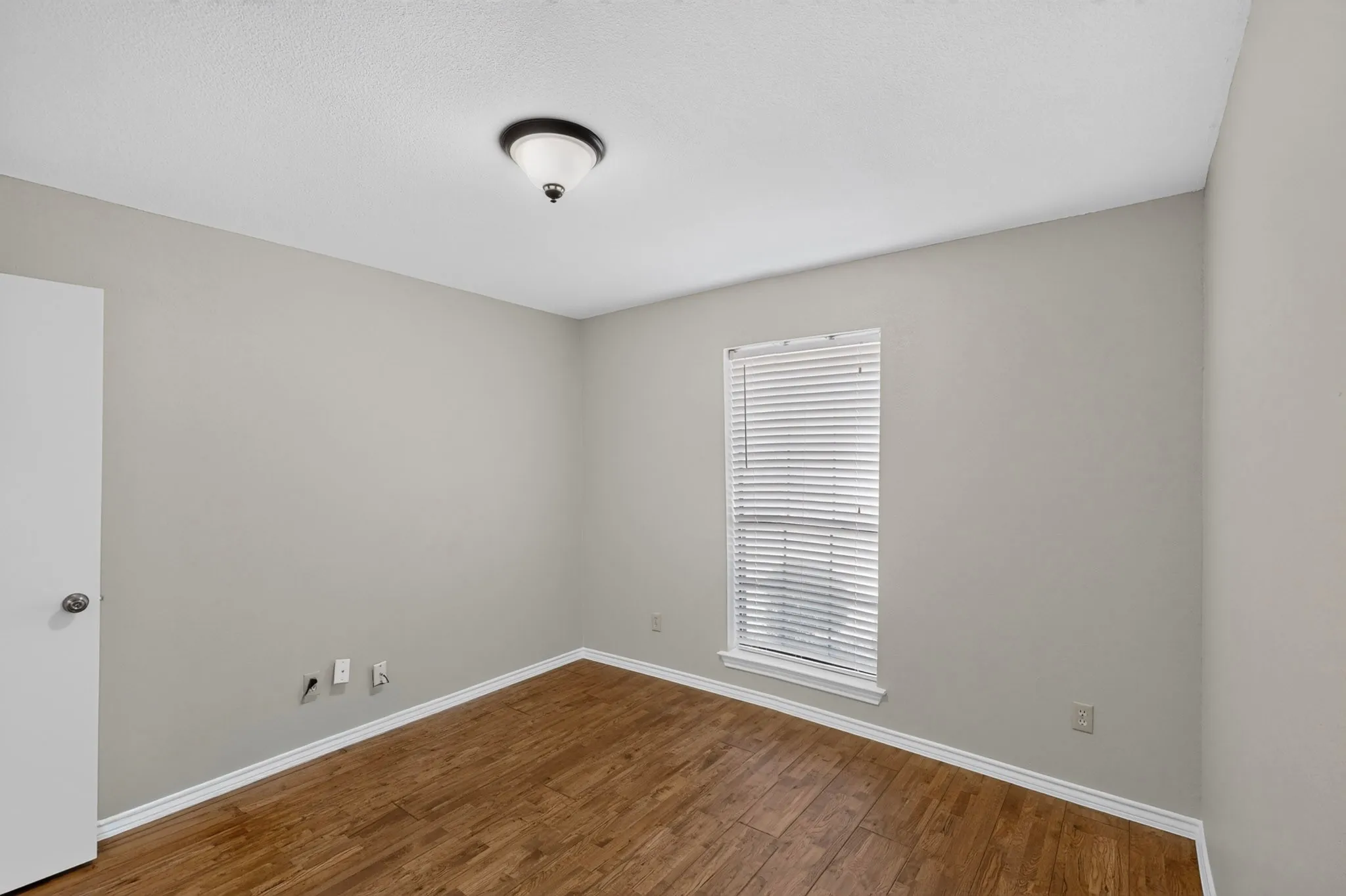 Bright bedroom showcasing warm wood-finish flooring, neutral wall finishes, and abundant natural light from a large window with plantation shutters
