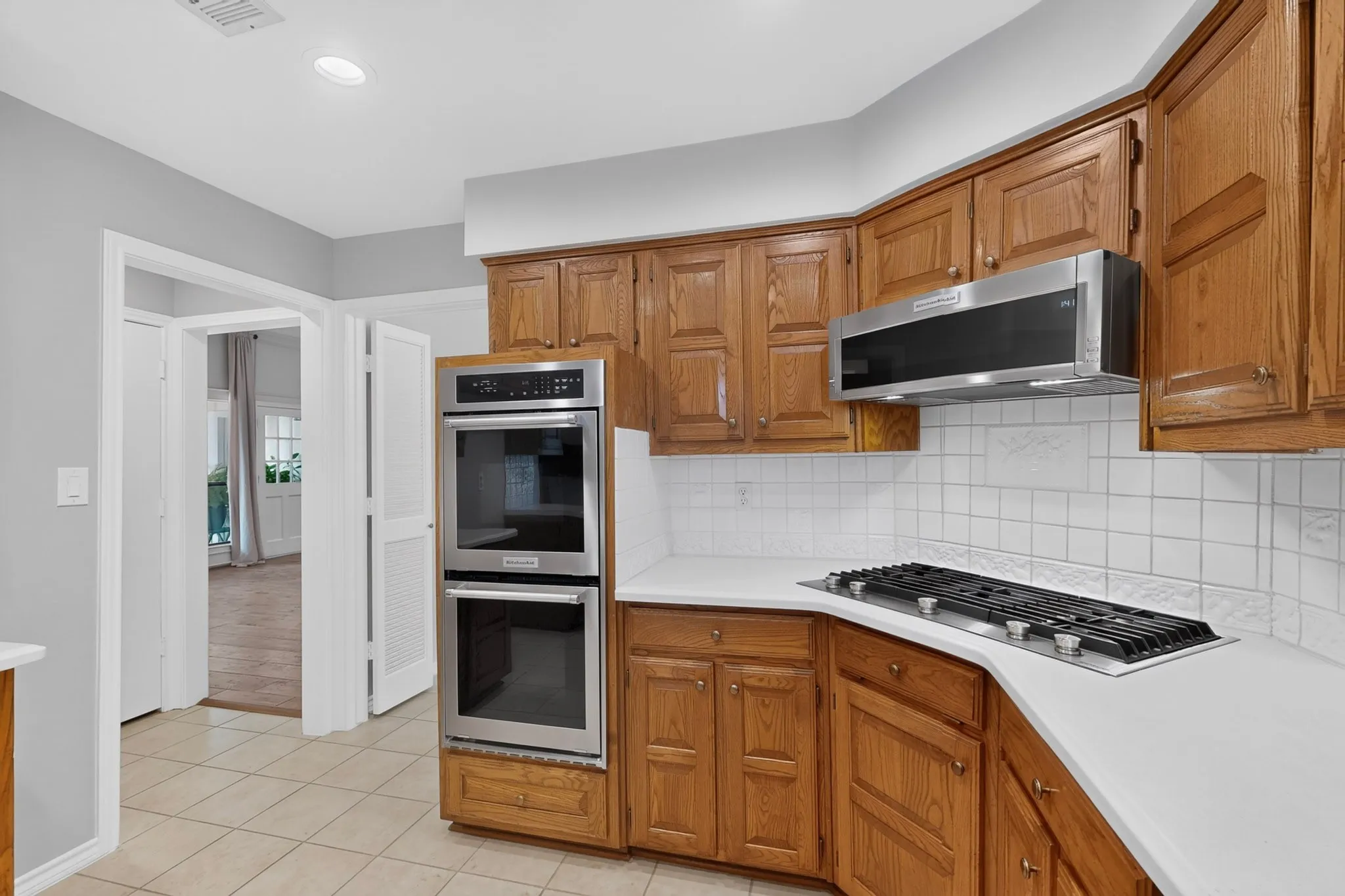 Chef's kitchen featuring warm wood cabinetry, stainless steel dual wall ovens, gas cooktop with black range hood, white subway tile backsplash, and tile flooring throughout the open layout