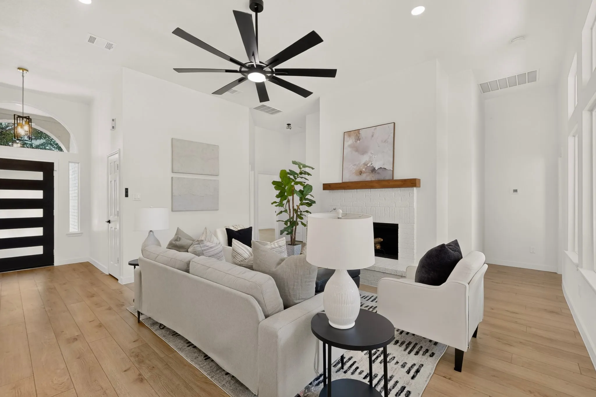 Wood-finish flooring throughout, modern black ceiling fan, white brick fireplace with wood mantel, arched entryway with transom window, and a black front door with horizontal lites