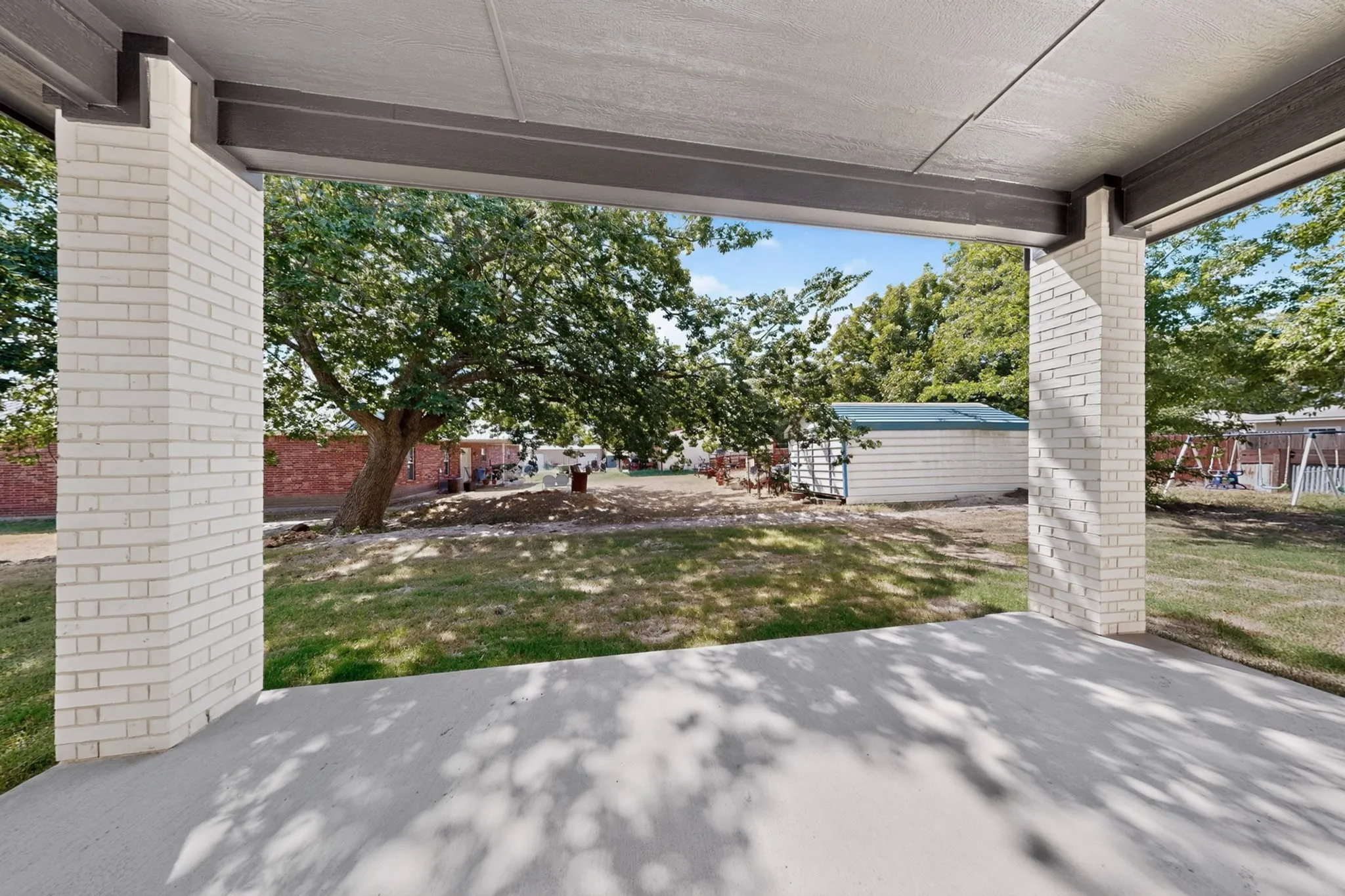 Covered patio with light-colored brick columns, concrete flooring, and a spacious green lawn