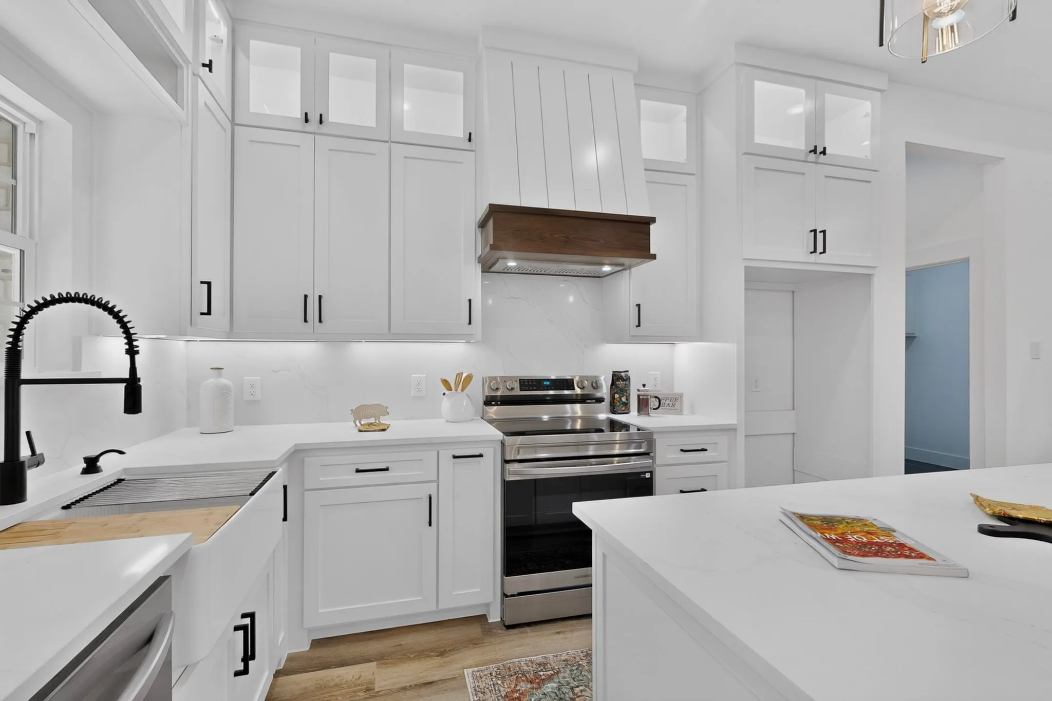 White shaker-style cabinetry with matte black hardware, stainless steel appliances, a farmhouse sink with a black coiled faucet, light countertops, and wood-finish flooring