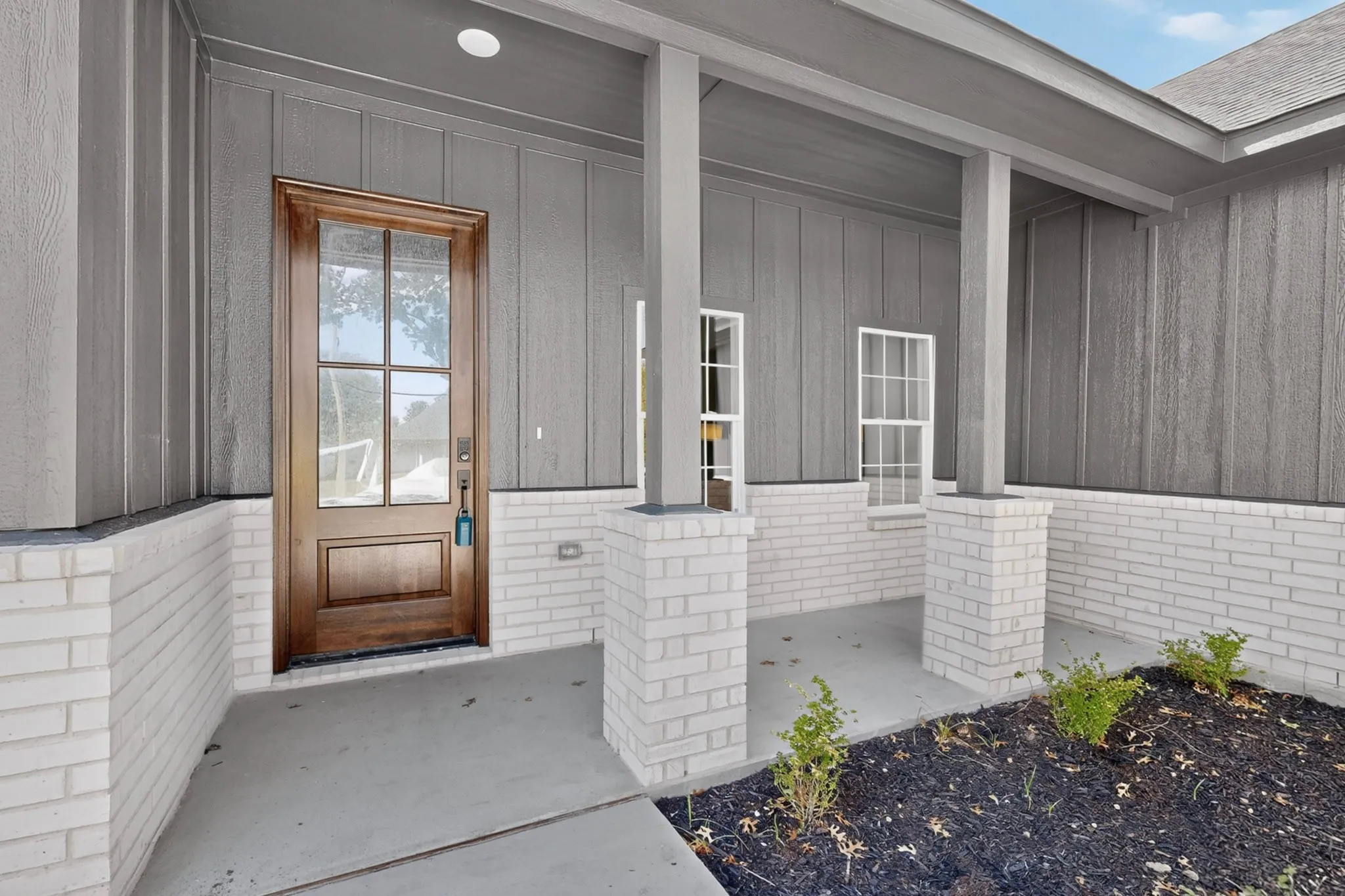 Covered porch featuring white brick columns and base, wood-paneled front door, and gray vertical siding