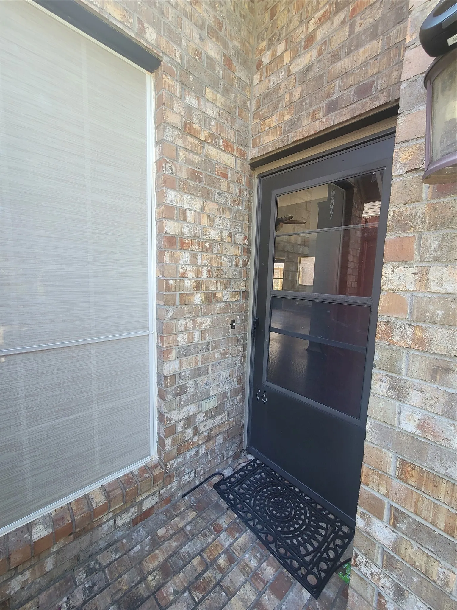 Brick exterior with a black screen door, an exterior wall-mounted light fixture, and a window with a sunshade