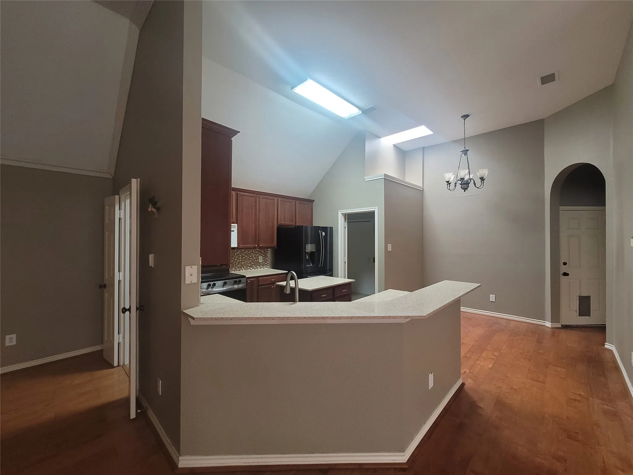 Open-concept kitchen and dining area featuring a curved island with white quartz countertop, stainless steel appliances, wood-finish flooring throughout, vaulted ceilings with skylights, and a decorative pendant chandelier