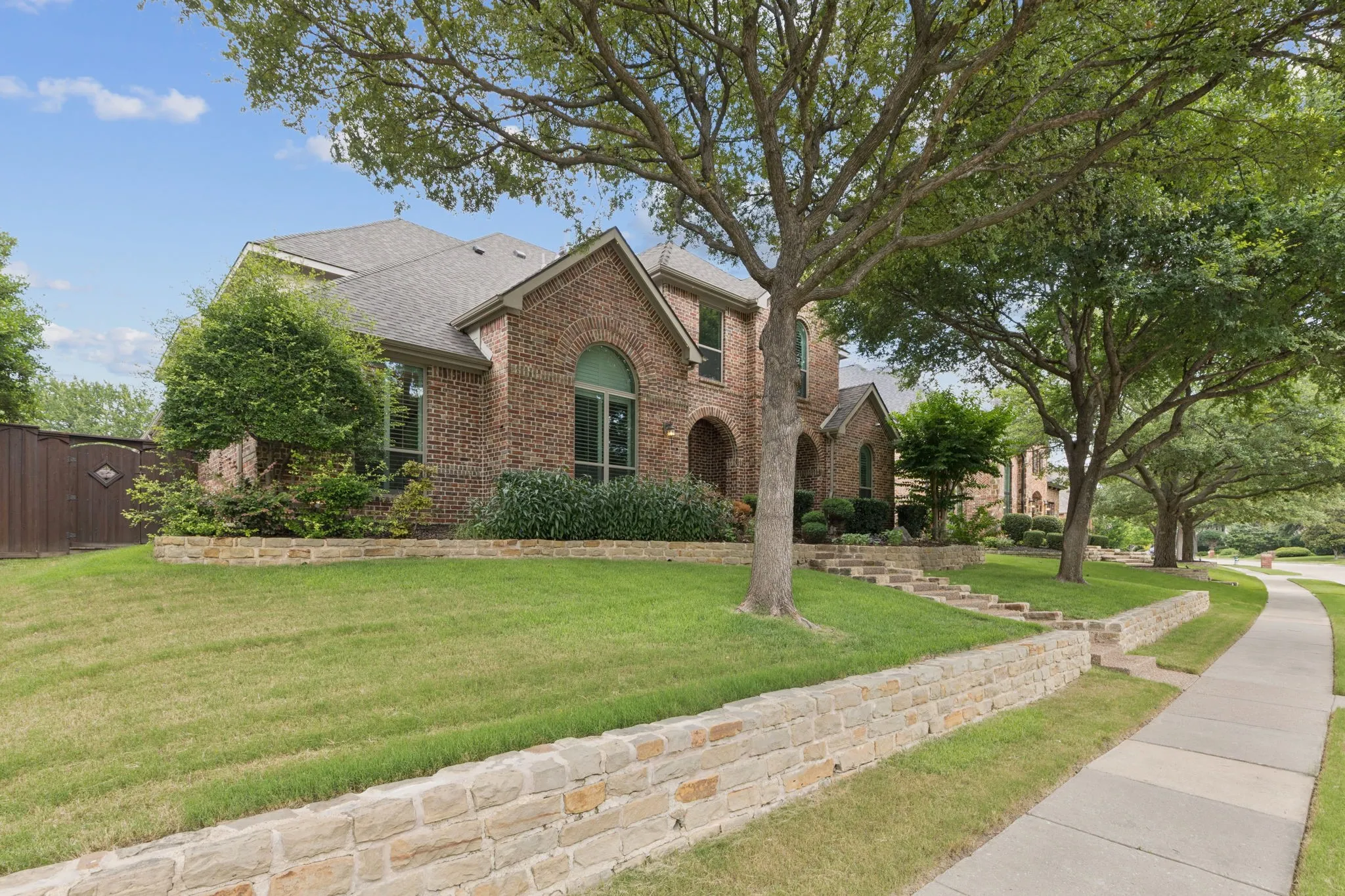 Brick facade with arched windows, tiered landscaping featuring stone retaining walls, mature trees, and a concrete sidewalk