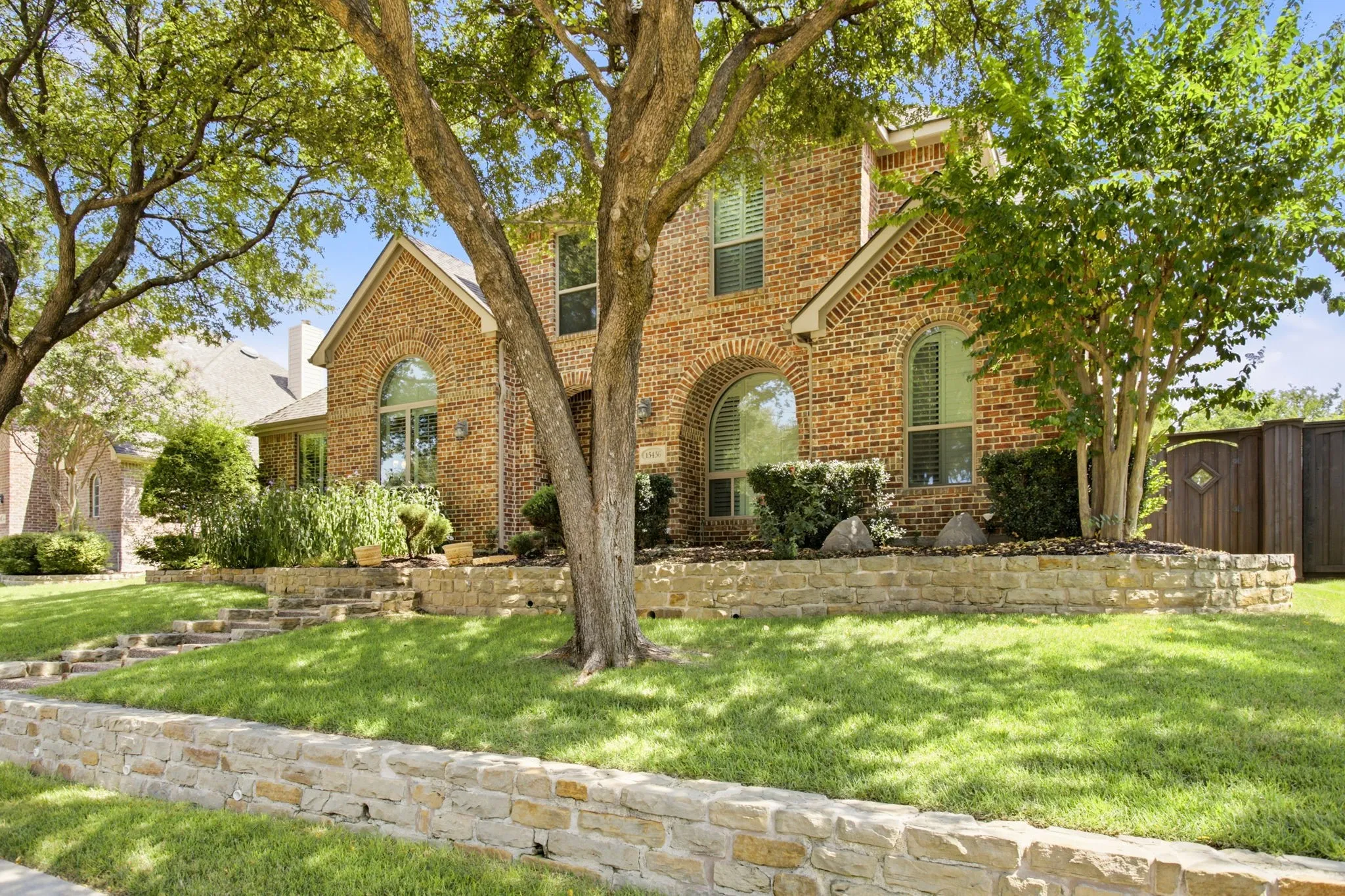 Brick exterior with arched windows, multiple gables, stone retaining walls, and mature trees