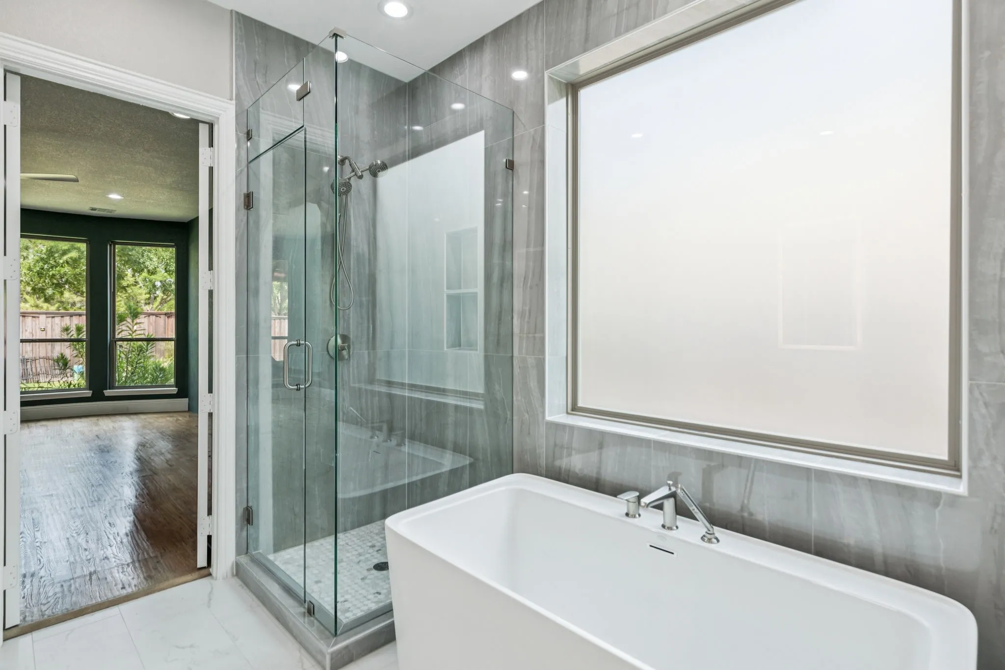 Spa-inspired primary bathroom featuring a freestanding soaking tub, frameless glass shower enclosure with tile surround, dual vanity with integrated sink, polished chrome fixtures, recessed ceiling lighting, and access to an outdoor garden space through French doors